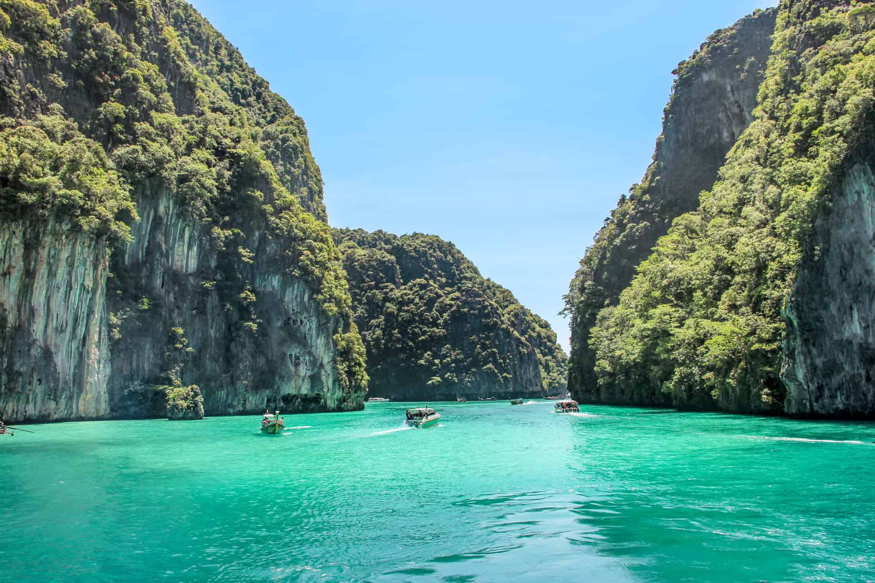 Boats glide through a wide stretch of emerald green waters between grass covered limestone rocks in Koh Phi Phi Leh in Thailand. 