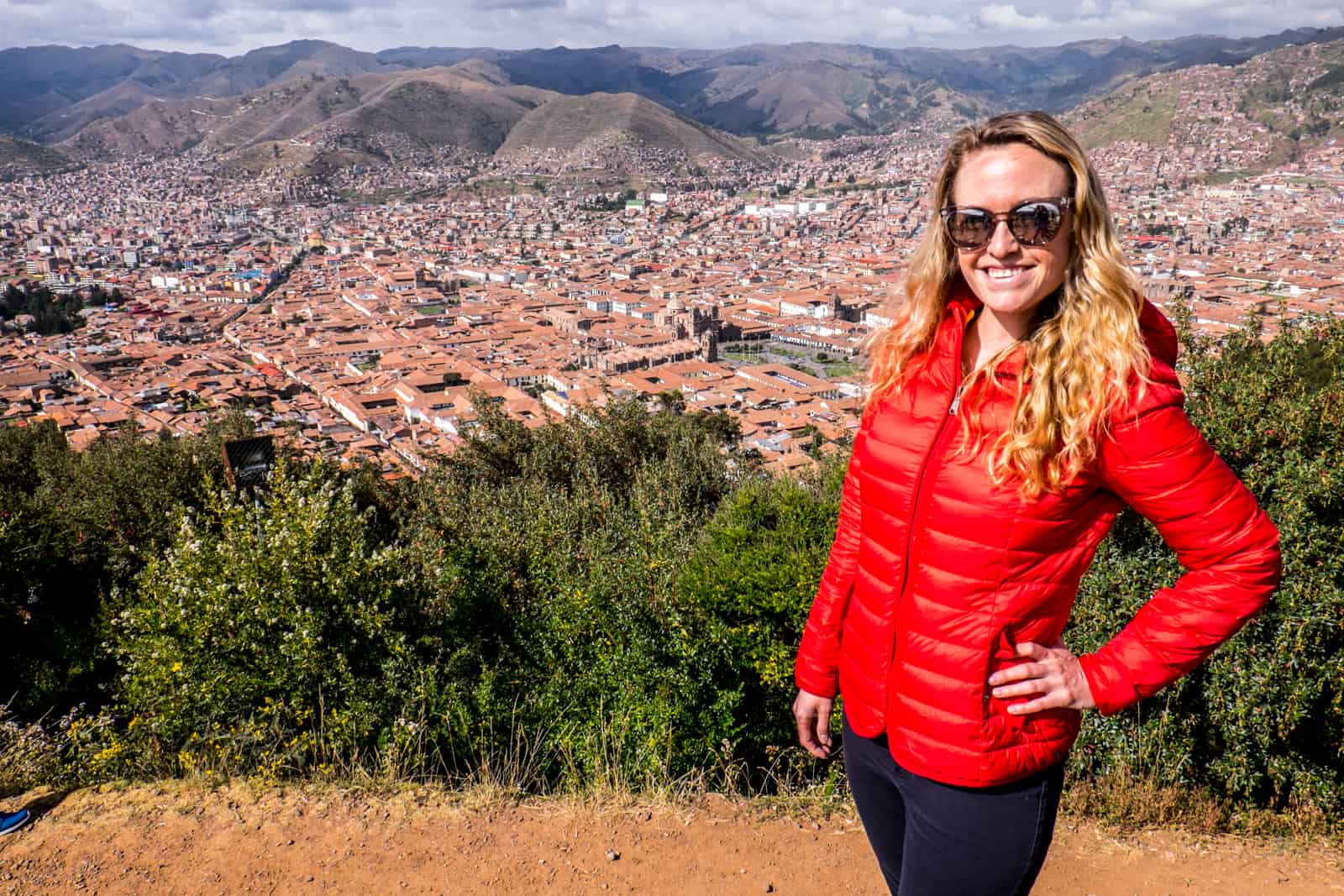 A woman in a red down jacket standing on a elevated dusty road overlooking the mountainous valley of Cusco. 