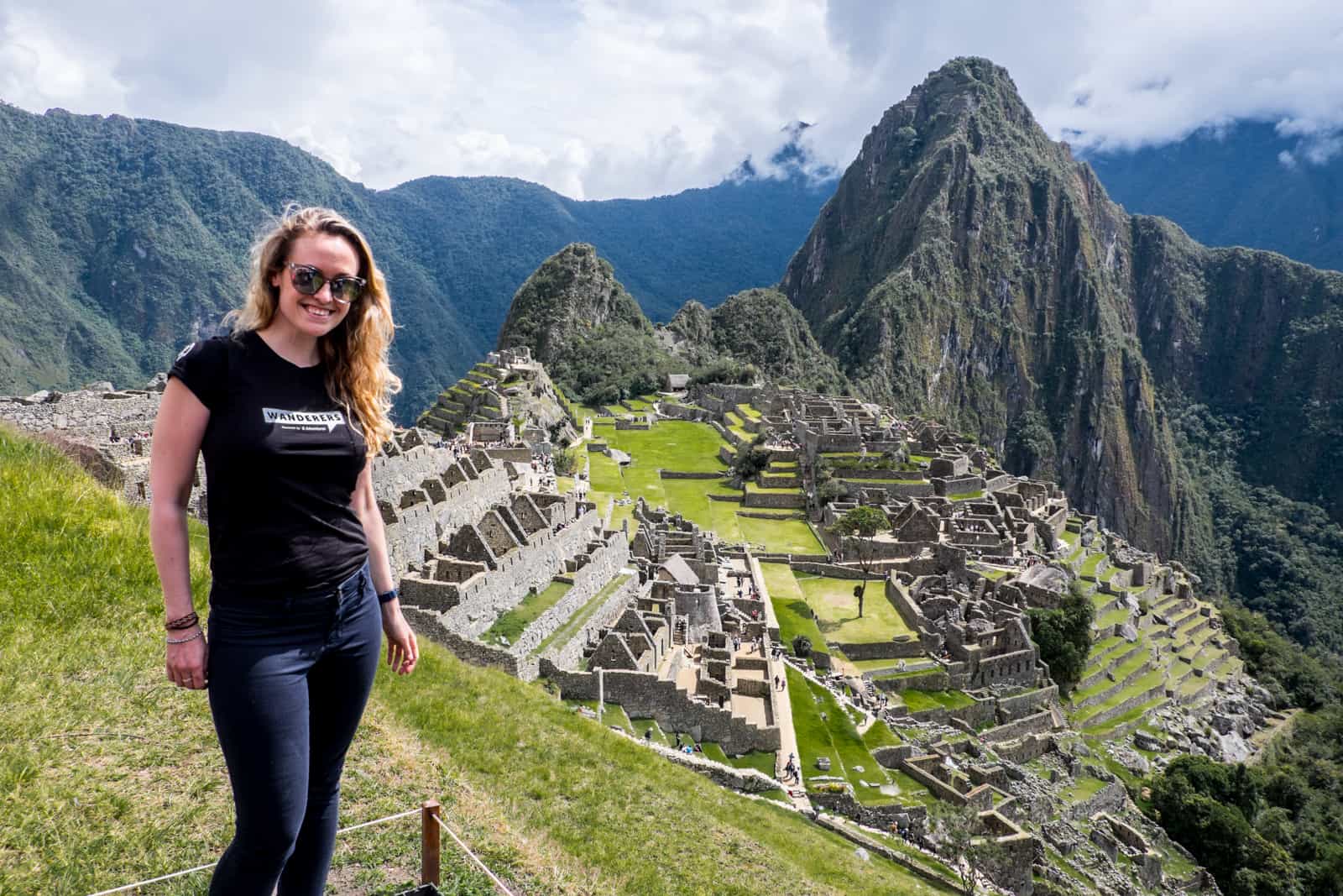 A woman stands on a grassy platform overlooking the stone ruins of Machu Picchu in Peru.