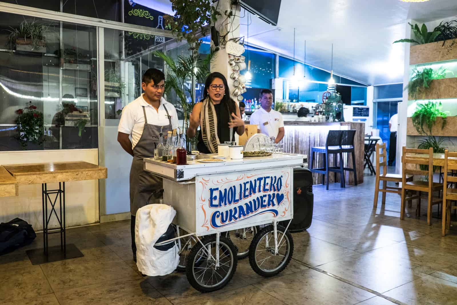 The owner of Nunay Reimy restaurant in Cusco explains traditional food and drink of Peru with produce displayed on a white cart.