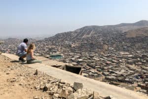 A man crouching and a woman sitting on a stone ledge overlooking a wide vista of a neighbourhood on a slope in Lima, Peru.
