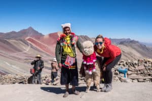 A tourist in Peru standing with a man and his alpaca at Rainbow Mountain (Vinicunca) in the Andes.