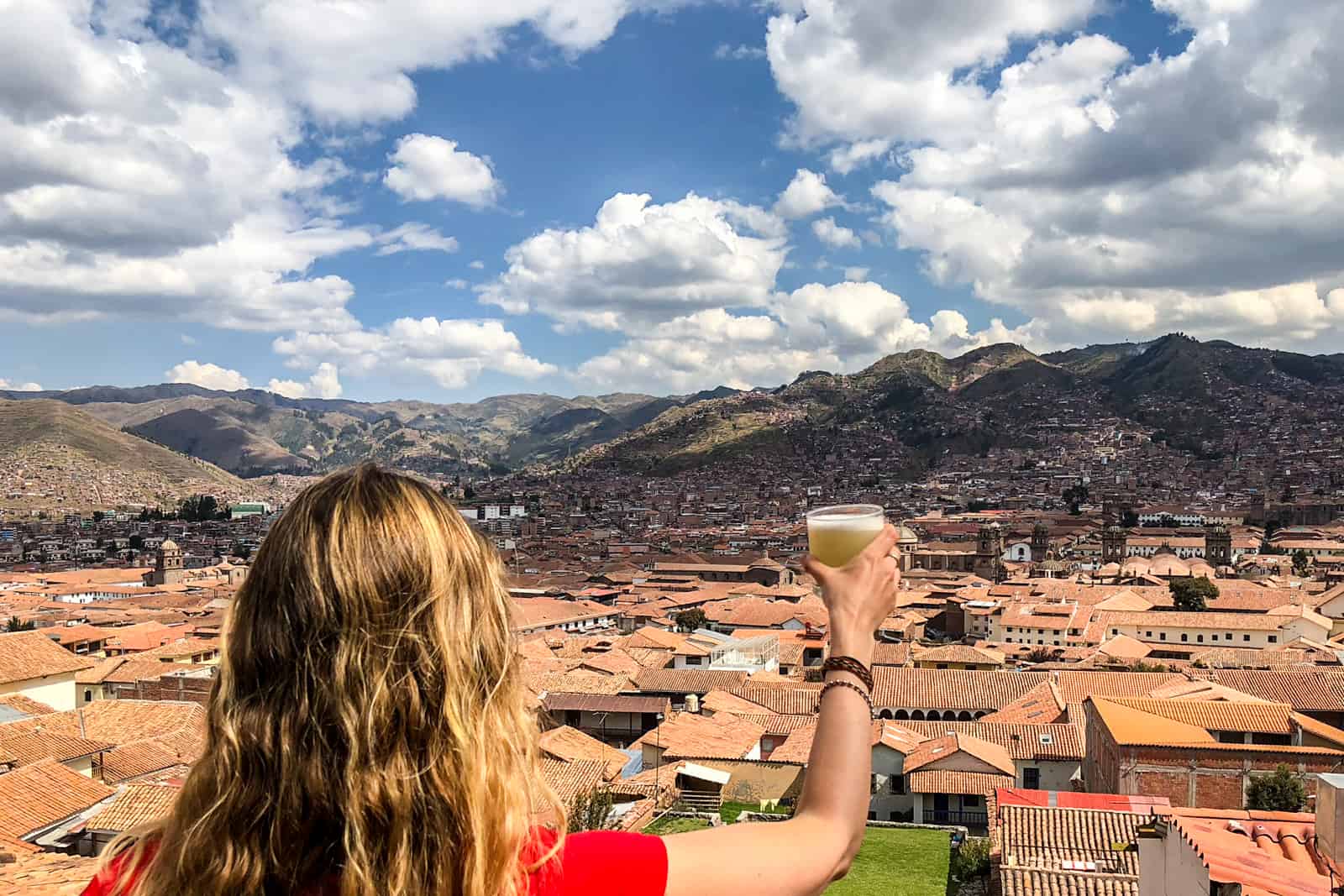 A woman raises a class of pisco sour overlooking a view of the red rooftops of Cusco valley backed by hills.