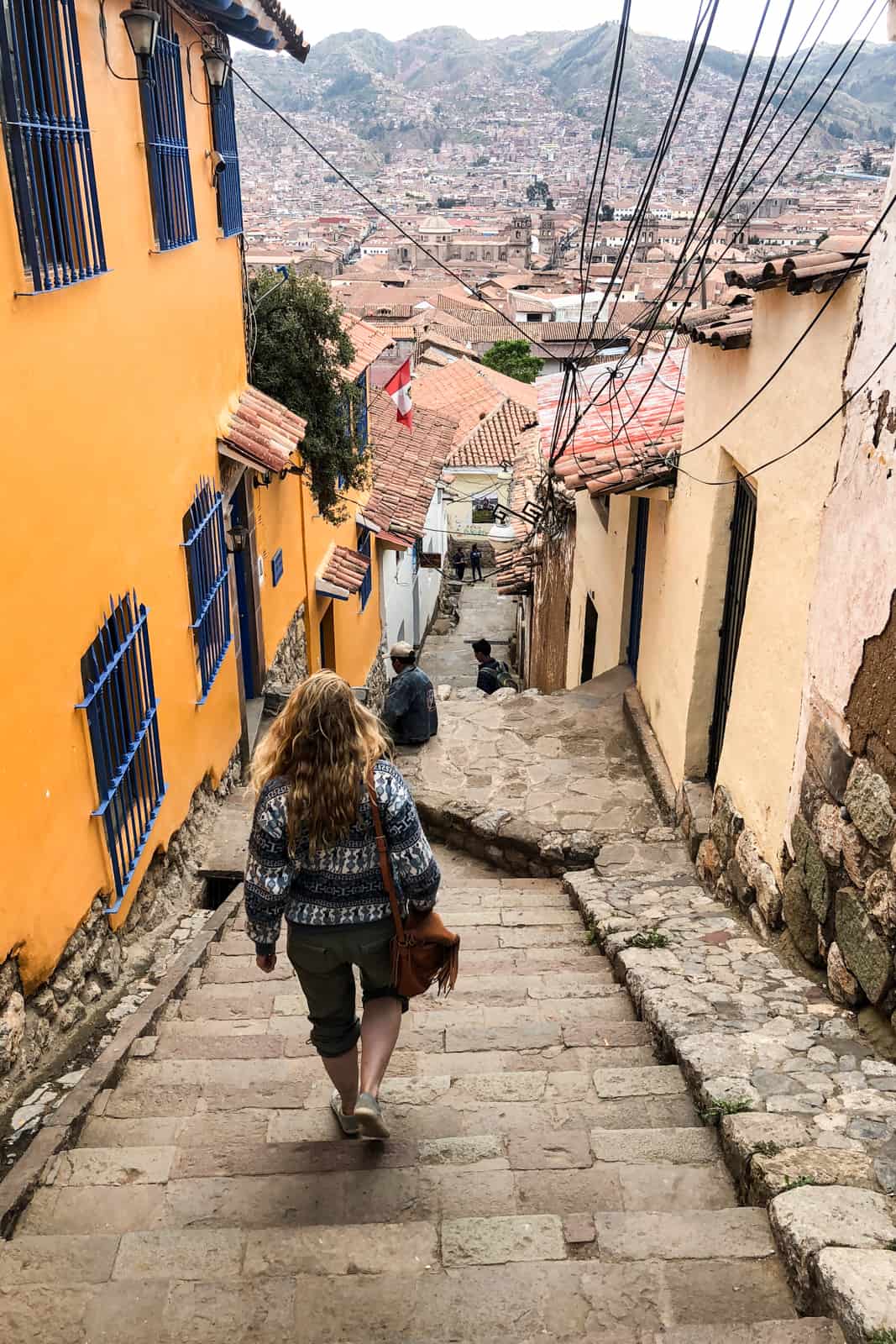 A woman walks down the stone steps of an elevated cobbled street of Cusco overlooking the valley. 