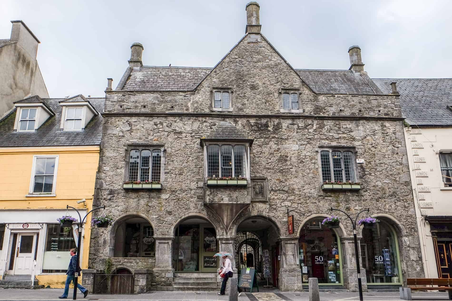 An old weathered stone house with triangular roof on a street in Kilkenny, wedged between more modern structures. 