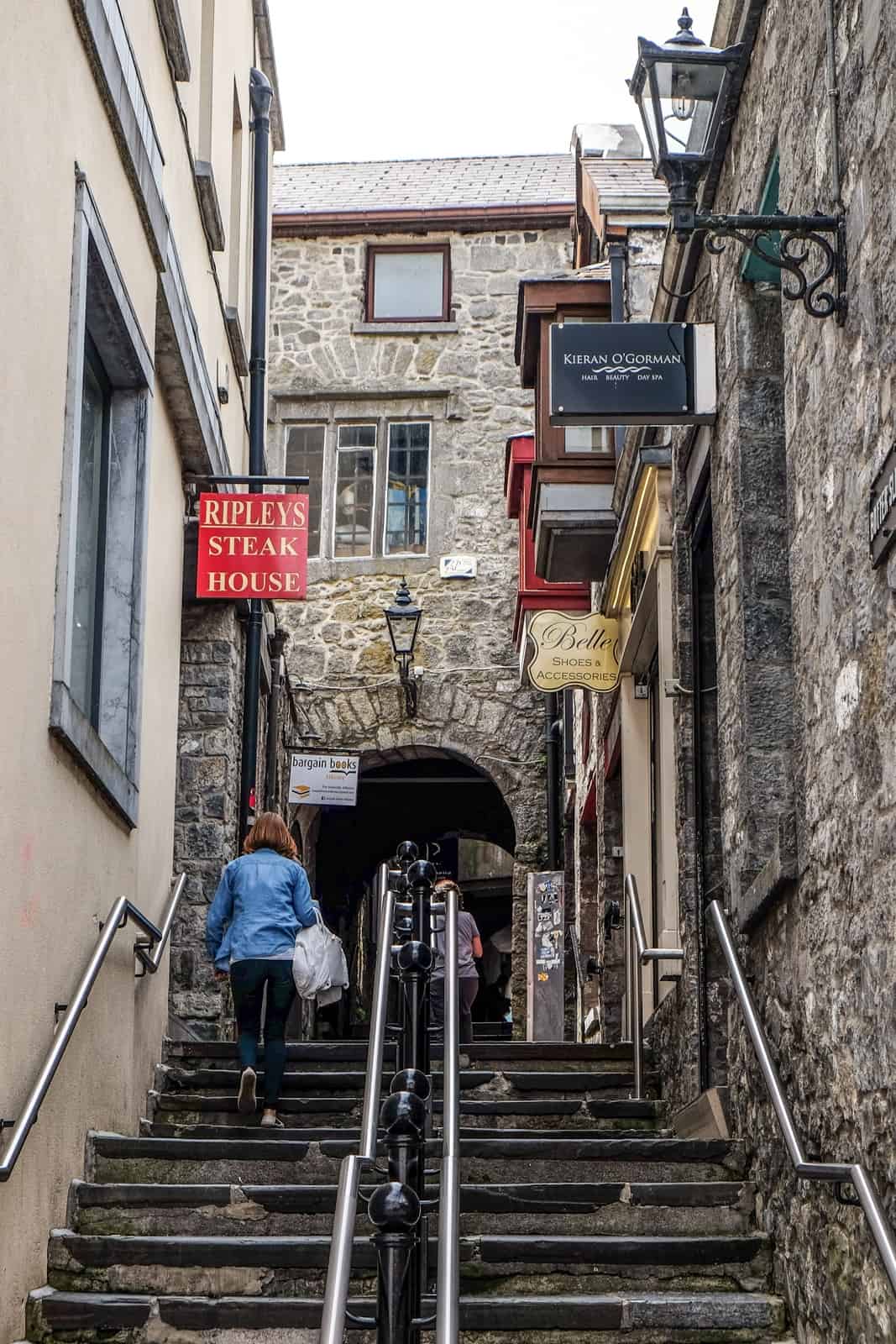 A woman walks up the stairs of a medieval street in Kilkenny surrounded by old grey stone buildings. 