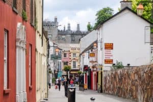 People walking down an old street in Kilkenny Ireland, filled with colourful painted houses, to the backdrop of a grey fortress building.