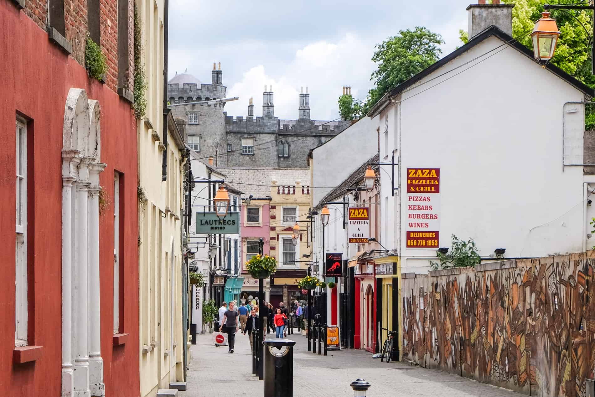 People walking down an old street in Kilkenny Ireland, filled with colourful painted houses, to the backdrop of a grey fortress building.