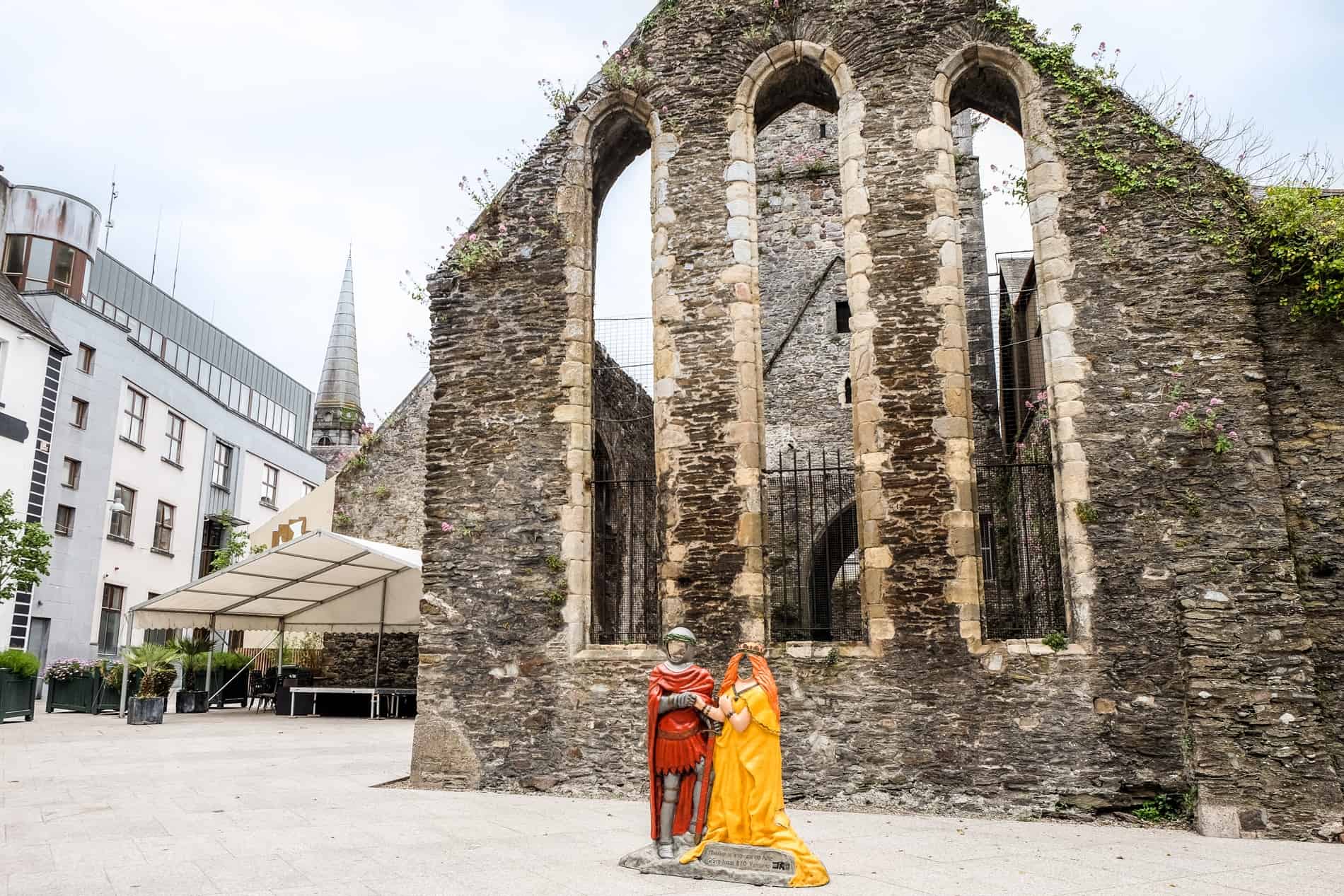 A red and yellow statue of Strongbow and Aoife in Waterford, standing in front of medieval ruins. 