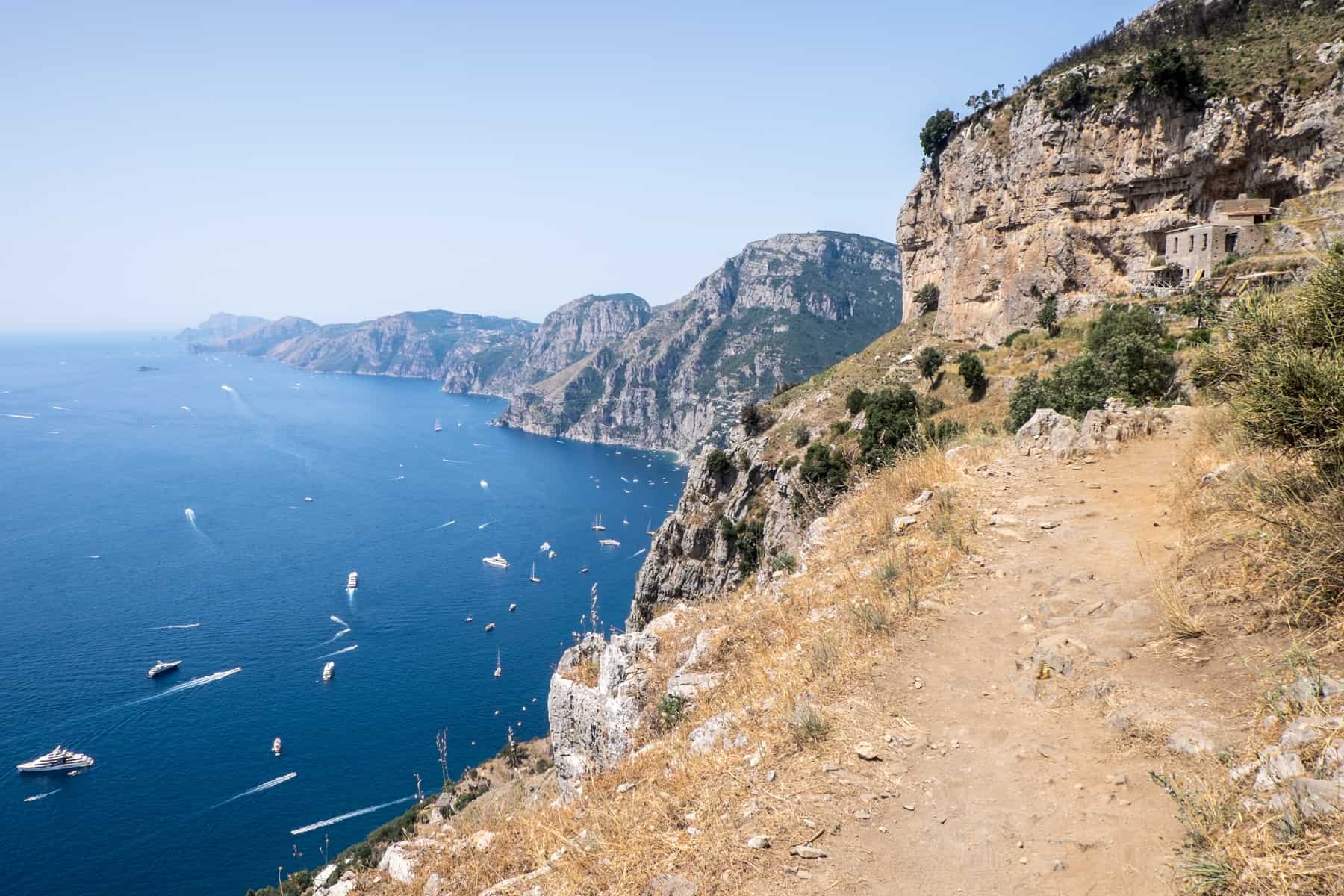 An elevated dusty, golden soil walking trail on the Path of the Gods hike looking towards a long stretch of mountainous coastline.