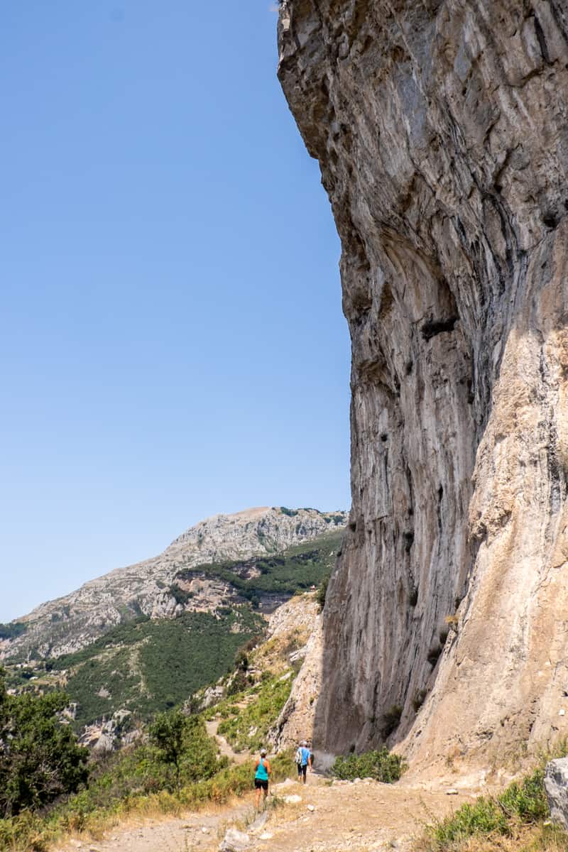 Hikers on the Path of the Gods walk past a high cliff face that curves outwards at the top, towards a small pocket of forest. 