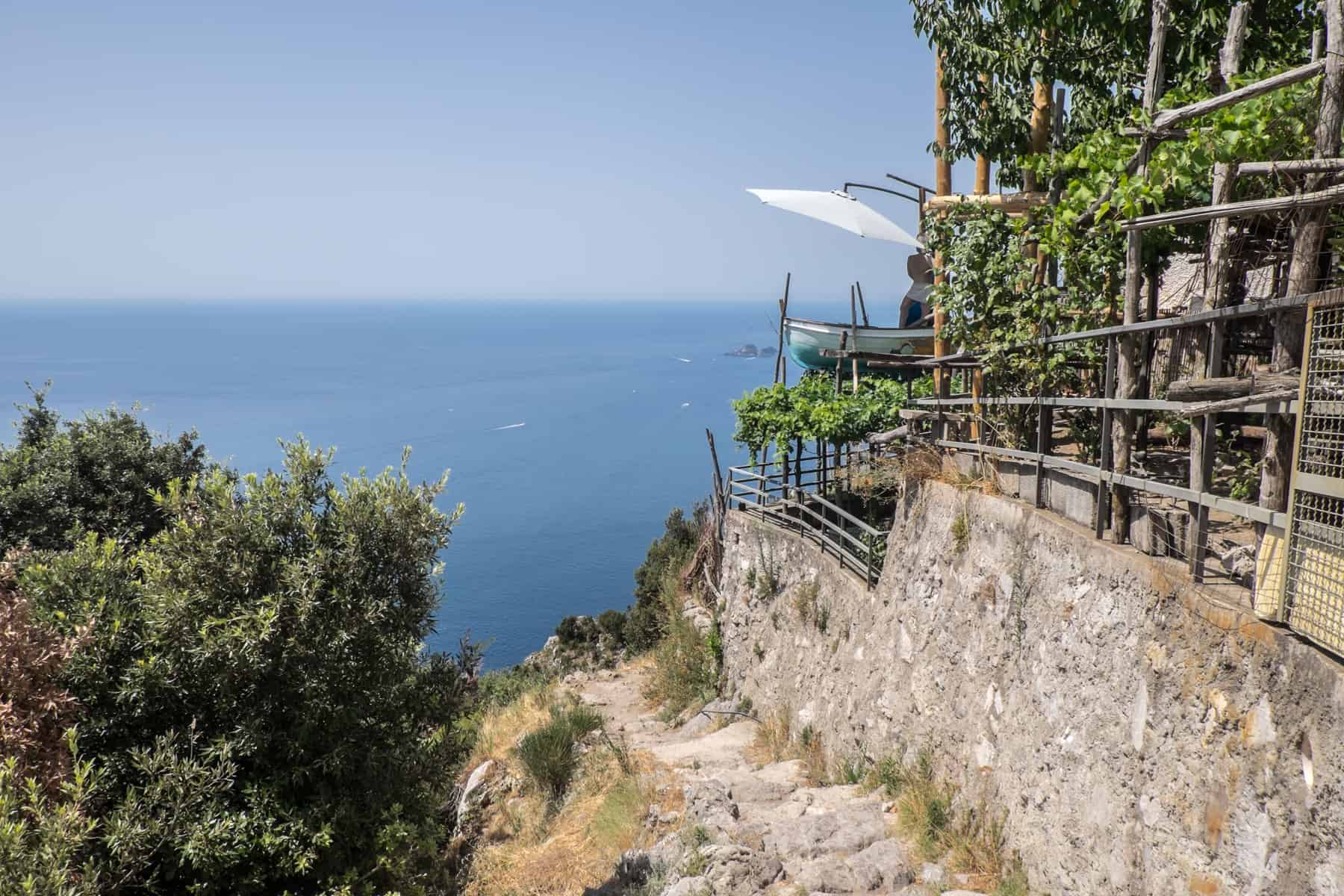 A rocky downhill path towards the sea next to a terraced cafe with a boat sticking out of the front. 