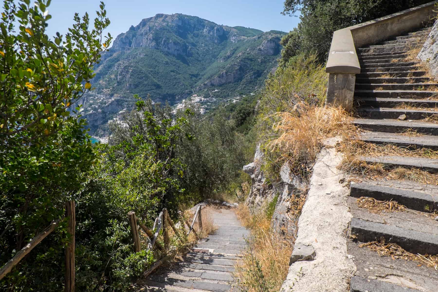 A long staircase, high in the mountains, curves and heads downwards towards Positano town seen in the distance. 