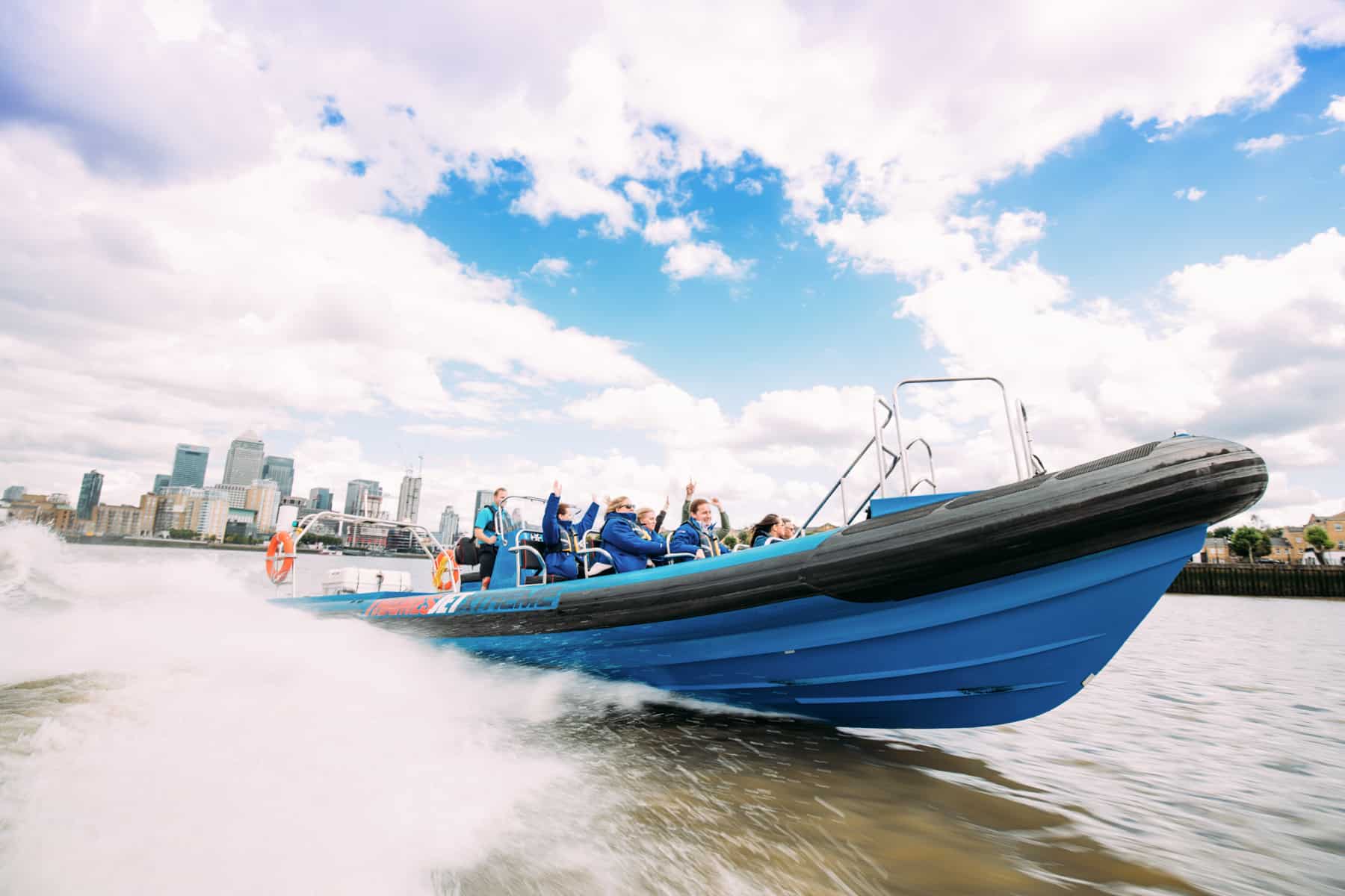 People with their hands in the air on a blue, high speed Thamesjet speedboat. Part of the London skyline can be seen in the background. 