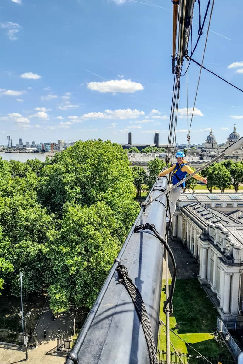 A woman wearing yellow and blue clothing hangs off the lower top sail of the Cutty Sark on the rig climb experience, with Greenwich in the background. 