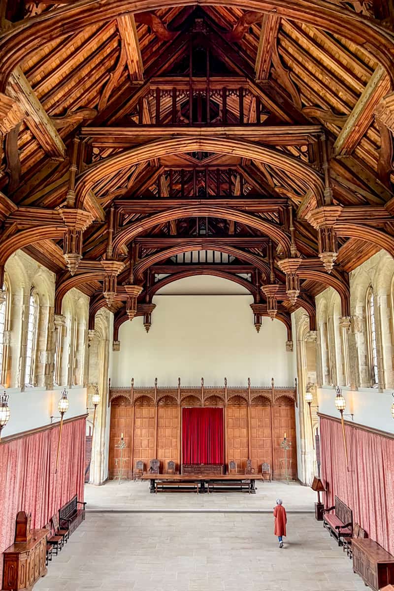 A woman in a red coat walks through the restored medieval Grand Hall at Eltham Palace with a thick wooden ceiling, high windows and red curtain trims. 