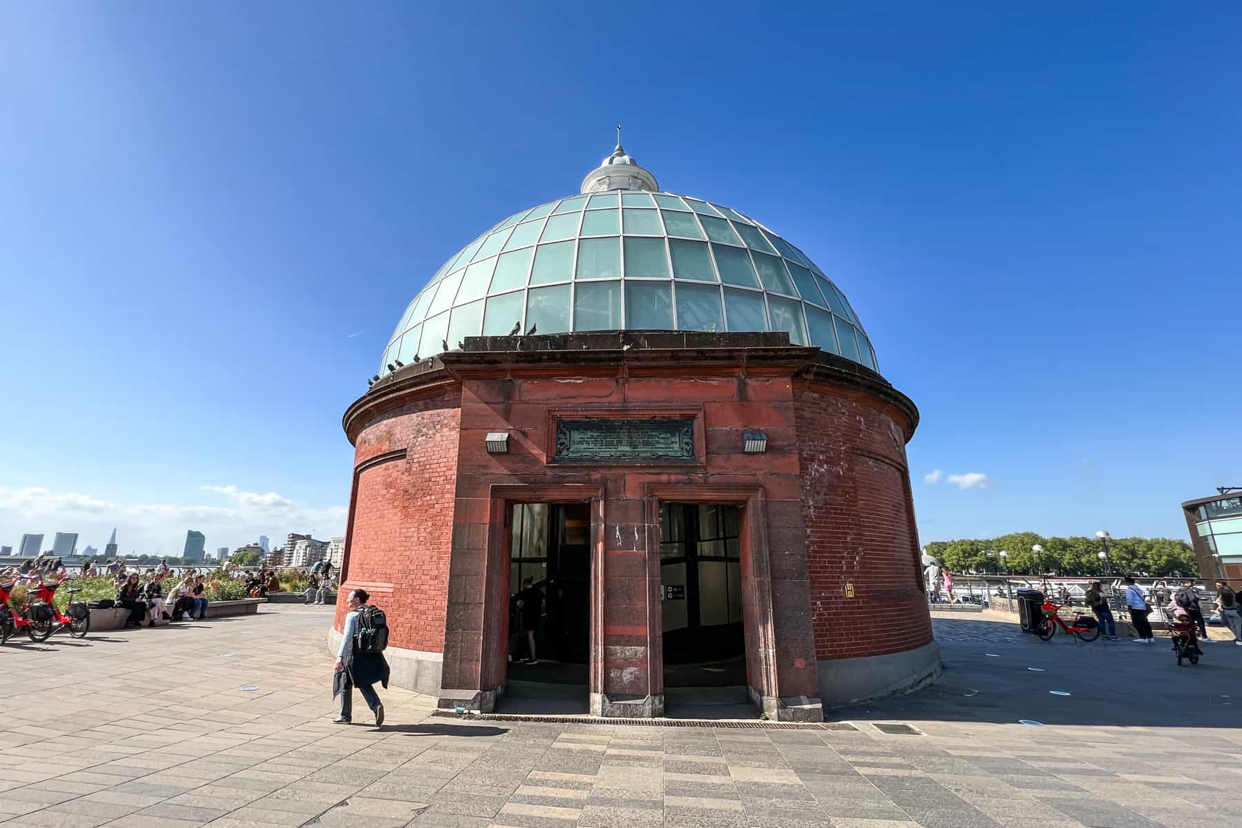 A round red brick building with a glass domed roof - the Greenwich Foot Tunnel Entrance - in front of the London skyline in the distance. 