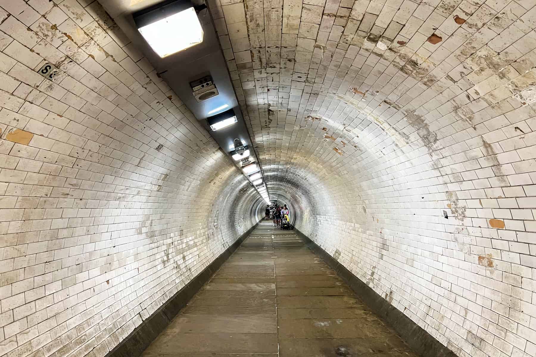 People walking through a long tunnel with white tiles and a row of ceiling lights - a crossing under the River Thames. 