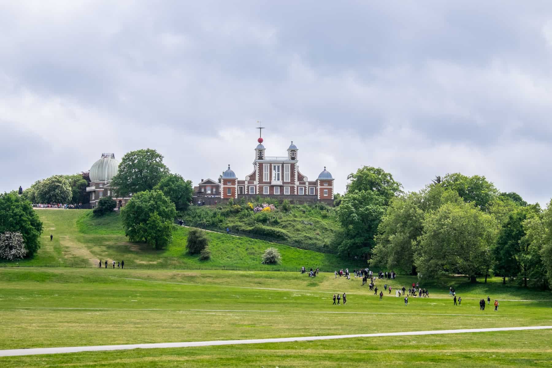 A red brick quad-spired observatory building at the top of a green park slope in Greenwich, London.