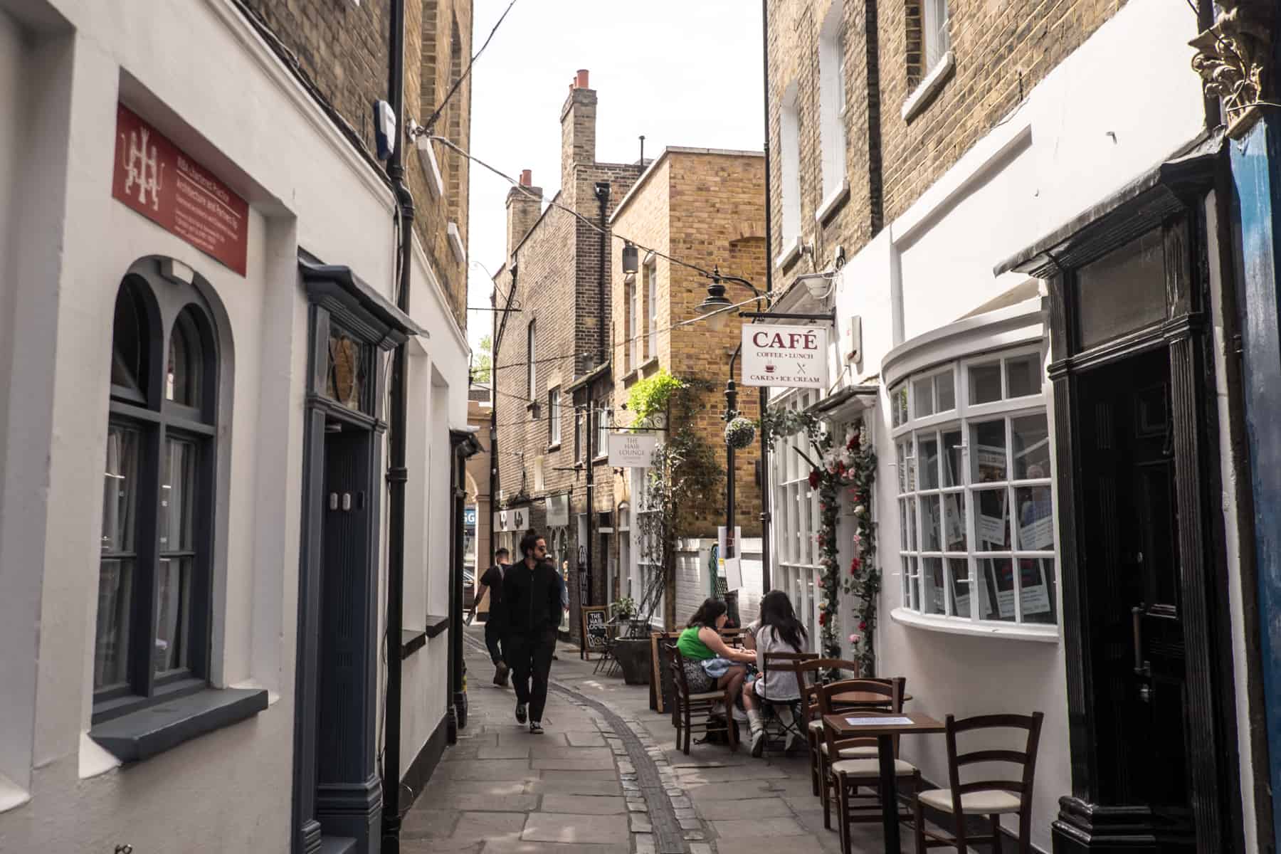 People walk through and sit outside a cafe on a narrow street in Greenwich with brick buildings and white shop fronts. 