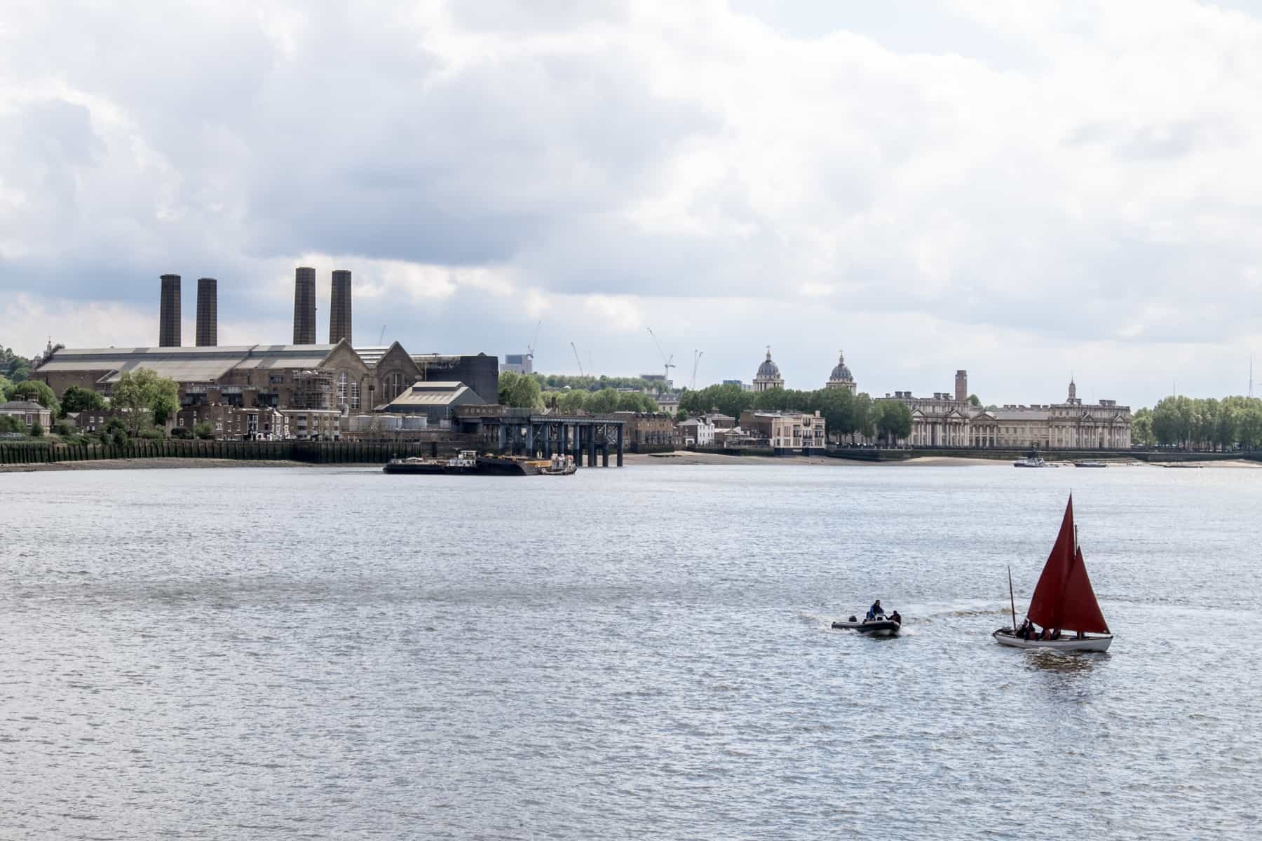 A red sail boat and small dingy boat on the River Thames in front of the coastline with Greenwich Power Station and Royal Greenwich domes and buildings. 
