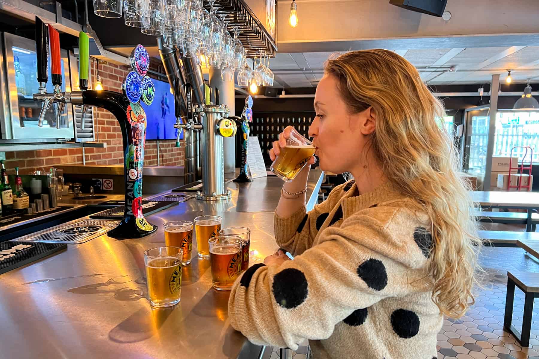 A woman with long hair and a beige jumper with black spots tries a selection of craft beers while standing at the bar. 