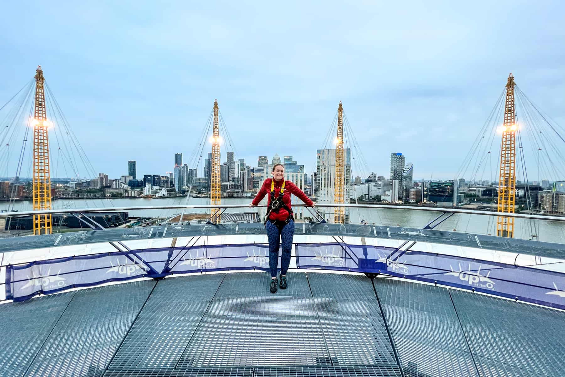 A woman in a red jacket, blue leggings and yellow harness stands on a platform on top of the O2 arena in London, to a backdrop of the modern city skyline. 