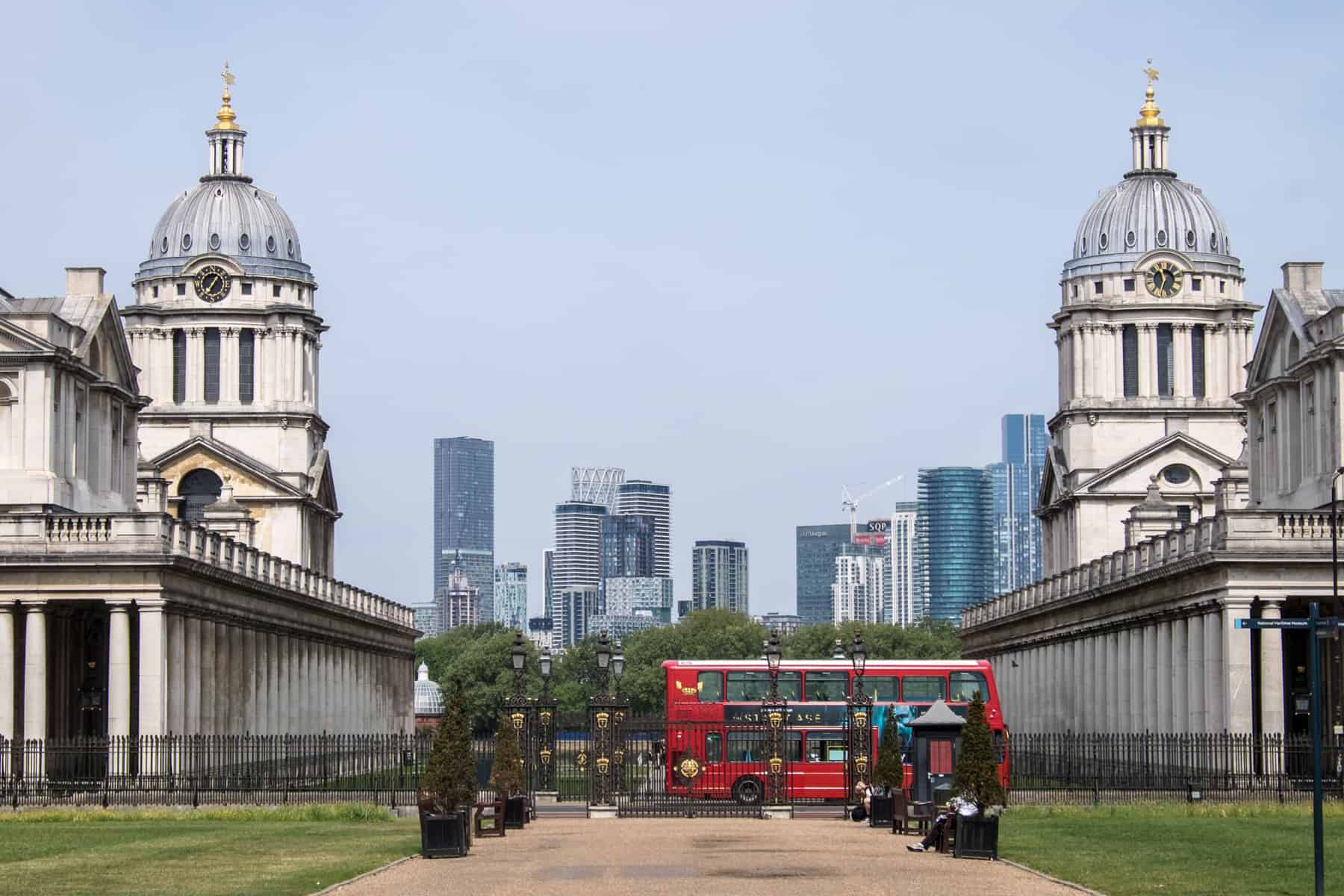 A red London buses drives between the two dome spires of the neo-classical Old Royal Naval College in Greenwich. Modern Canary Wharf can be seen in the background. 