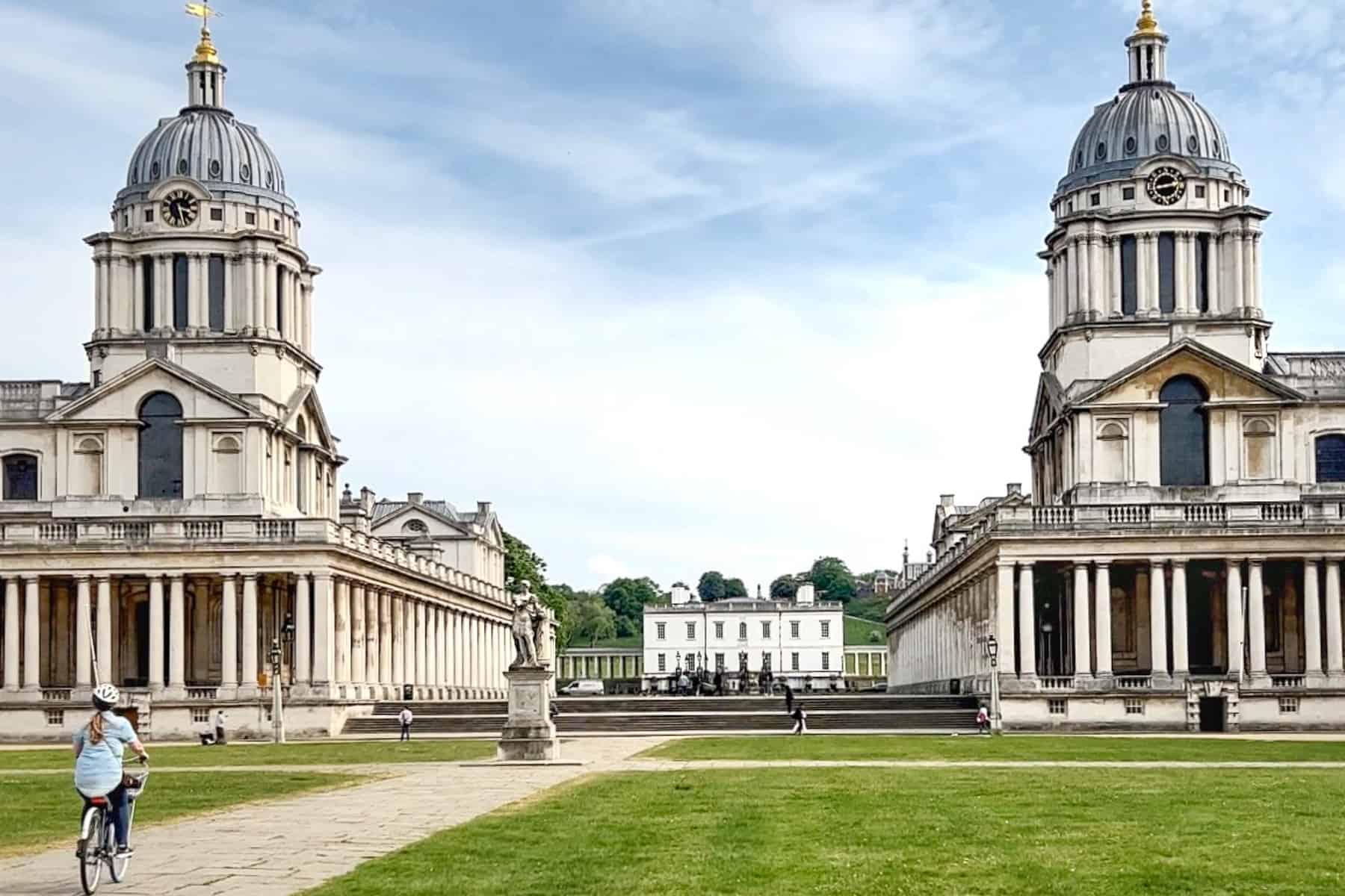 A woman on a bike cycling in front of the two-domed Old Royal Naval College and the Queen's House in between.