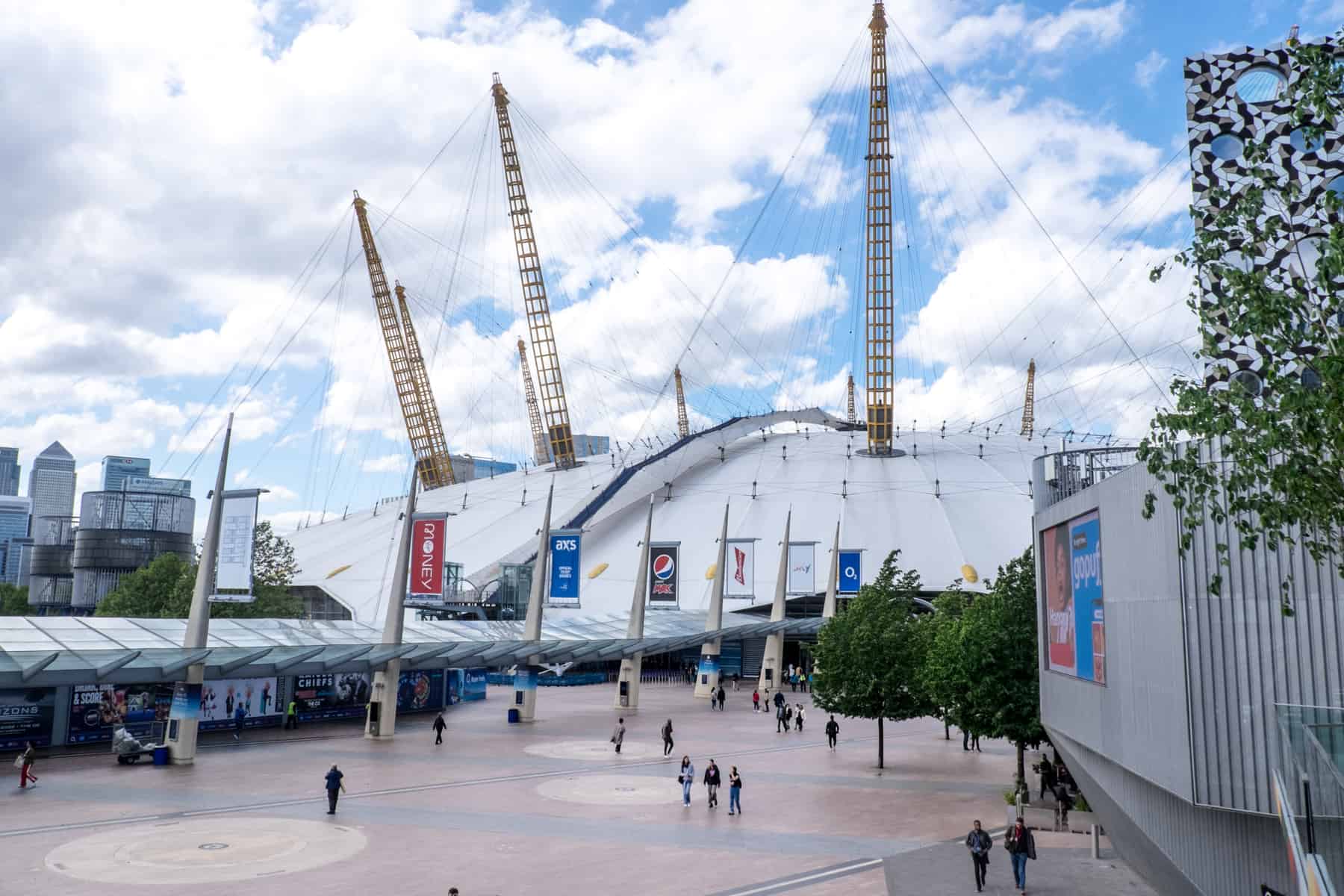 People walk in front of the O2 arena with a white dome exterior, yellow towers and a blue walkway over its roof. 