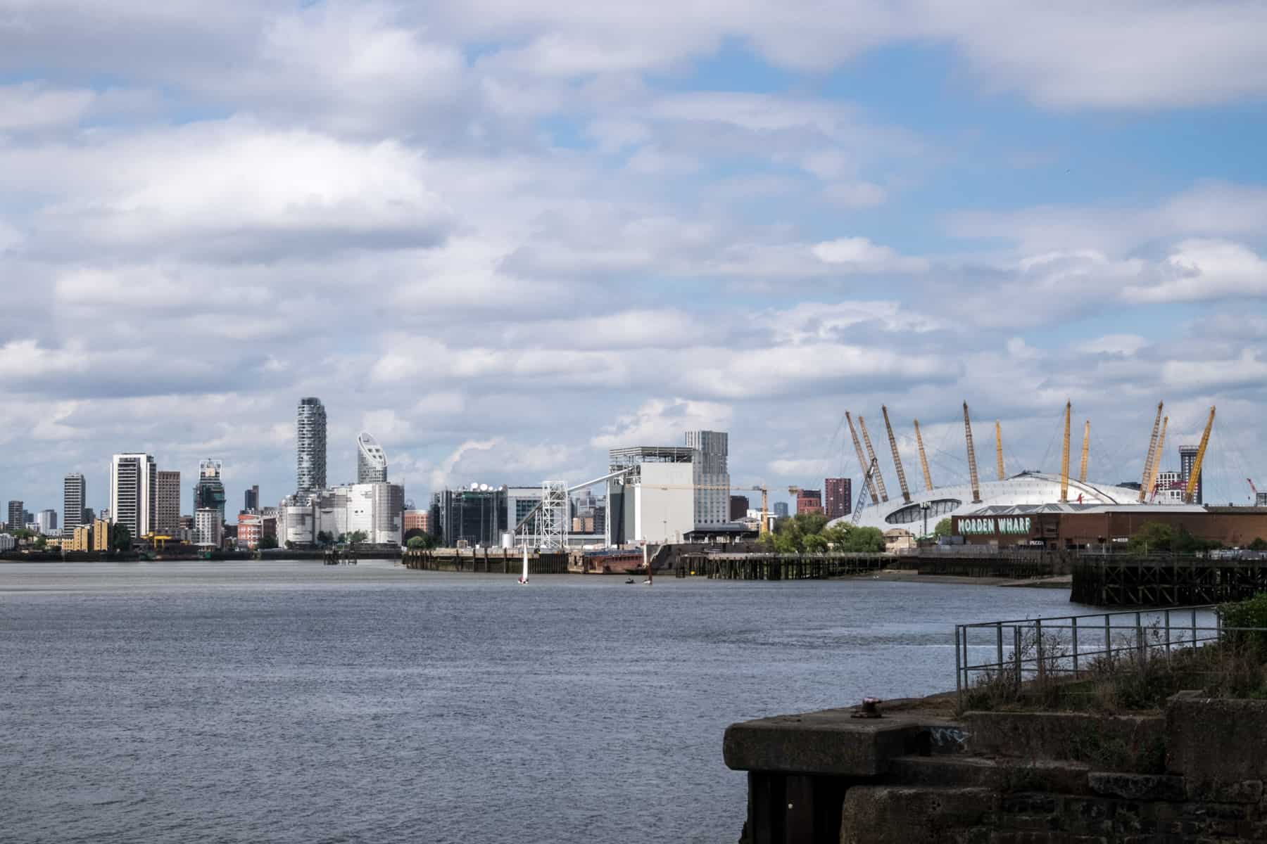 A view to the North Greenwich Peninsula from the Thames Path with modern high rise buildings and the white dome O2 Arena with yellow towers. 