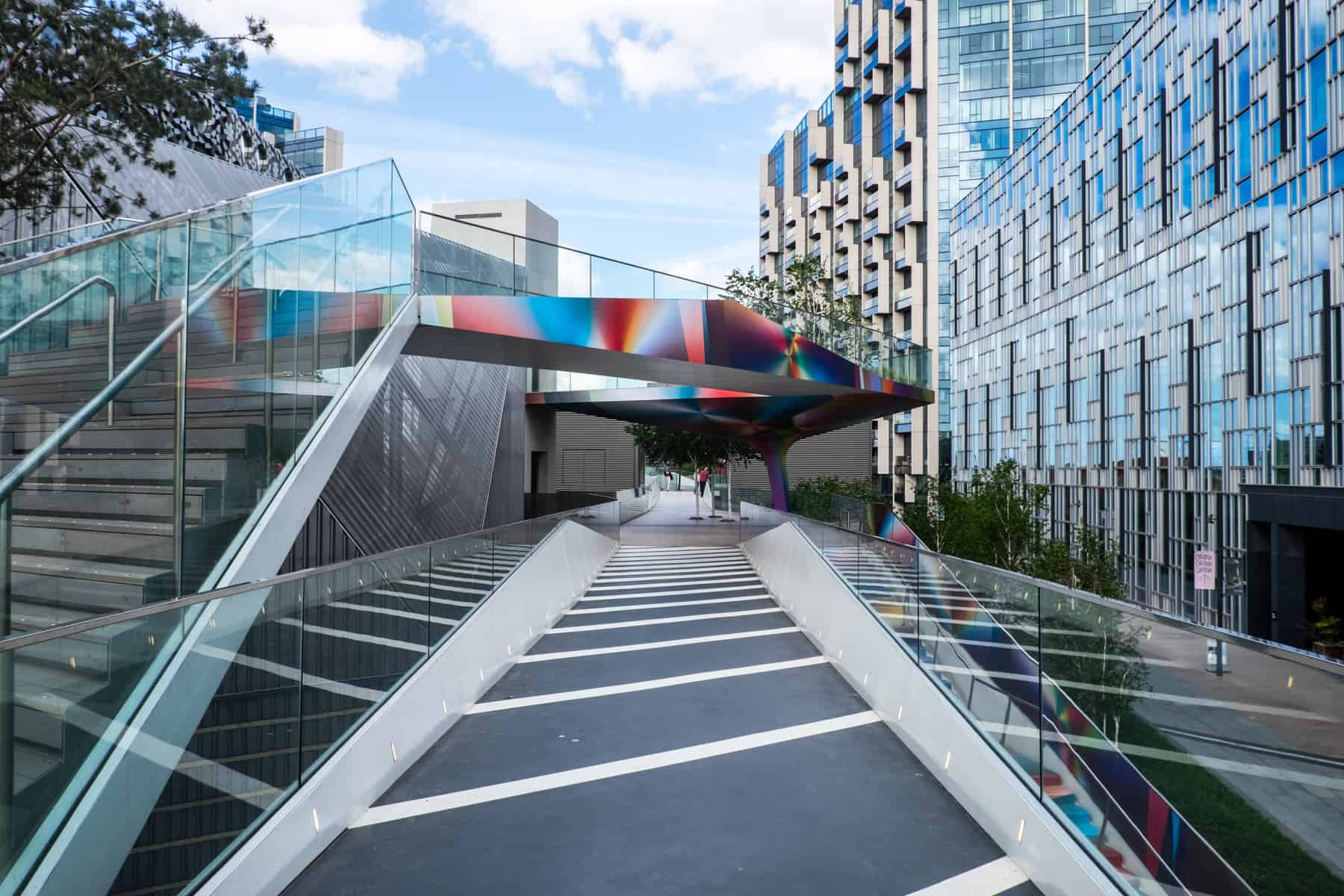 An art installation pathway in Greenwich Peninsula with rainbow balconies and monochrome walkways, next to modern glass buildings. 