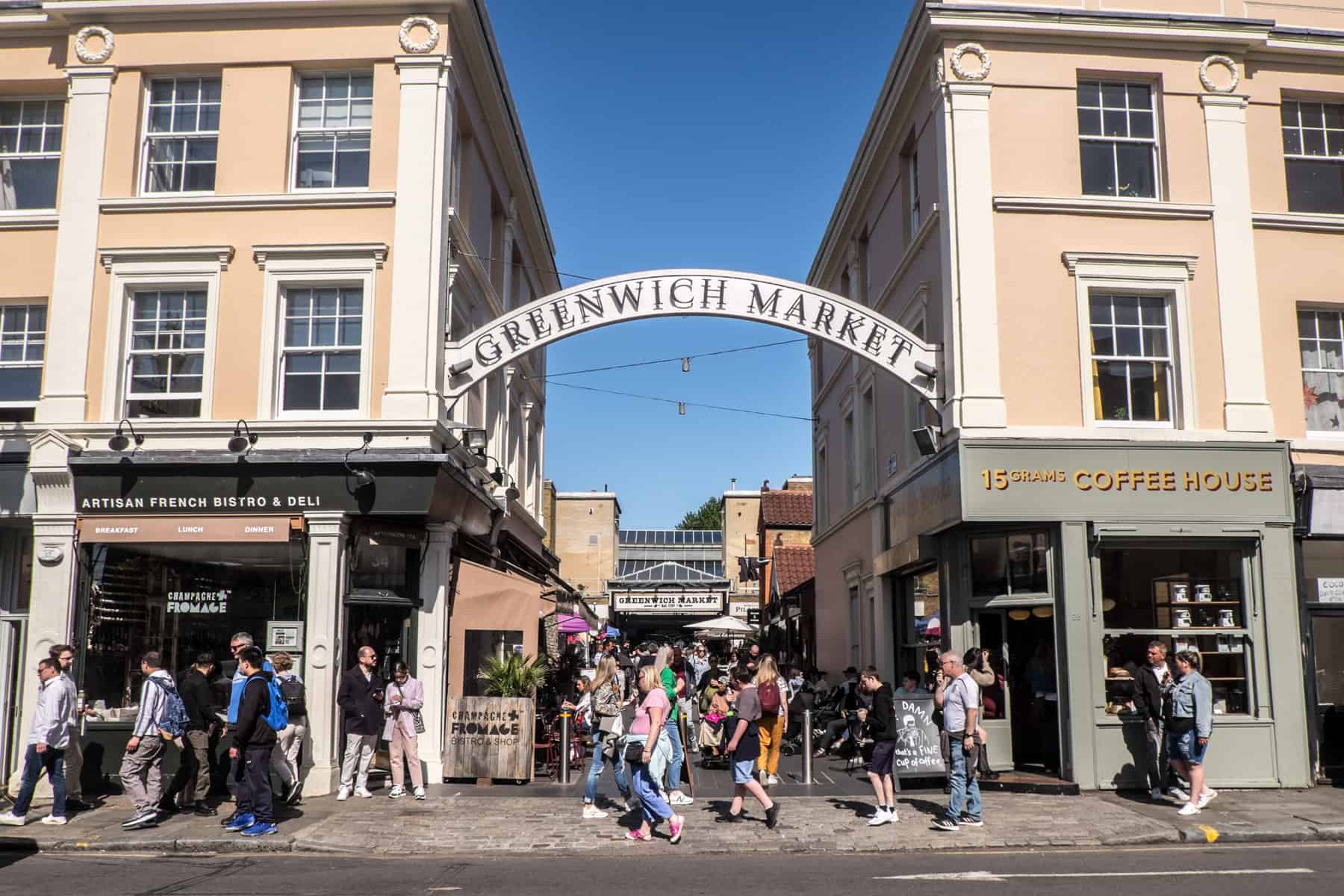 Crowds walk along the street outside the curved Greenwich Market sign and entrance. A Artisan French Bistro and 15 Grams Coffee House can be seen on either side. 