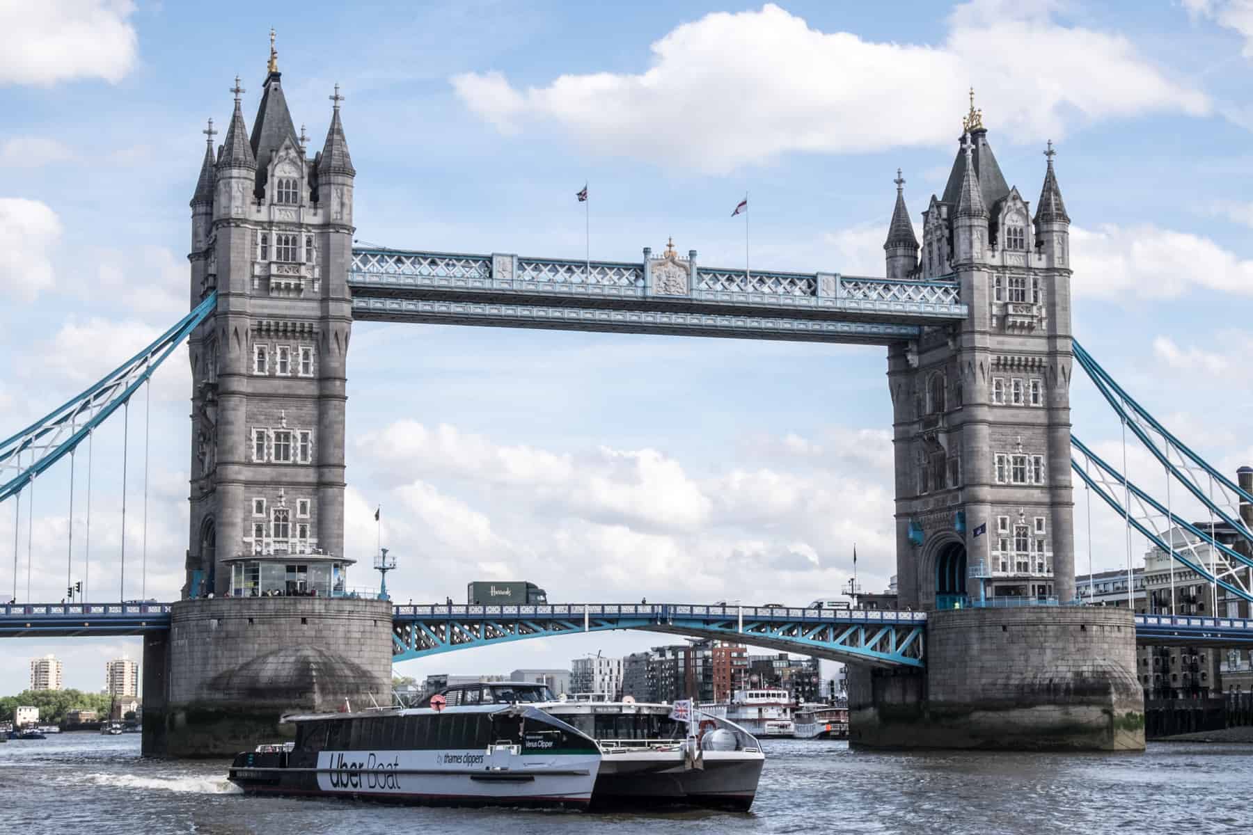 A black and white Uber boat passes the grey and blue Tower Bridge on the Thames. 