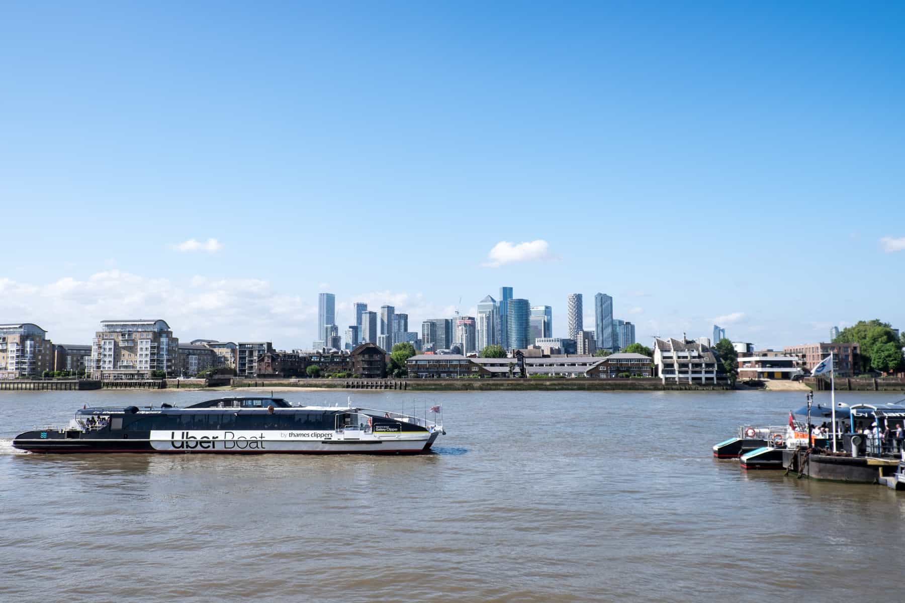 An Uber Boat by Thames Clippers driving towards a Pier on the River Thames with the Canary Wharf skyline in the background.