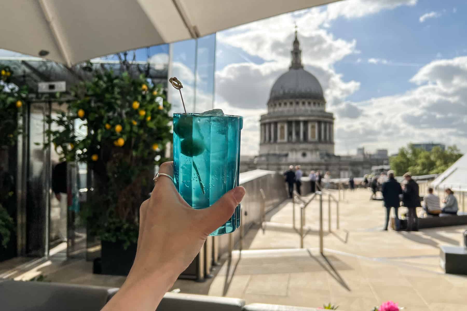A hand holding up a blue cocktail glass at a rooftop bar overlooking St. Paul's Cathedral in the background. 