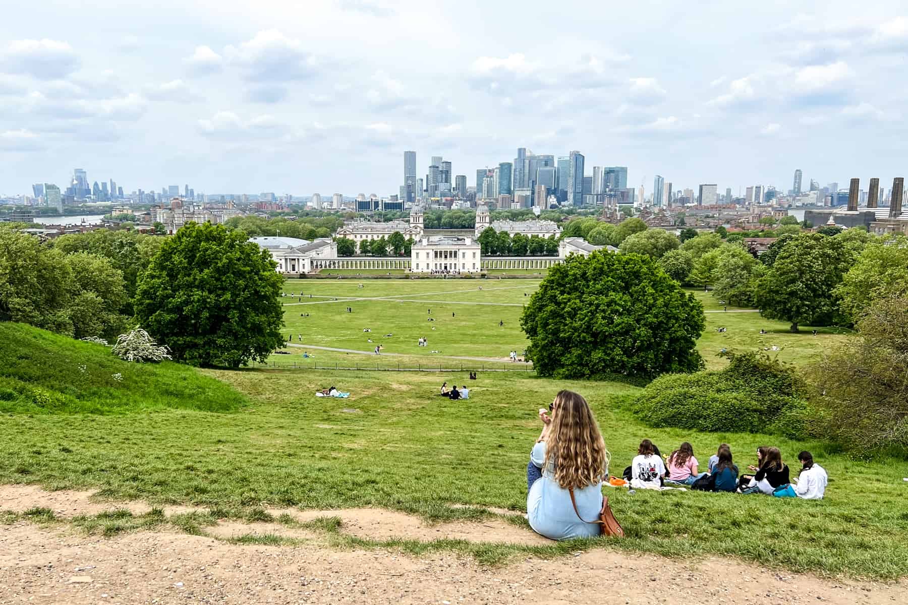 A woman sits on a hill overlooking Greenwich Park and the Royal Naval College complex and the City of London skyline.