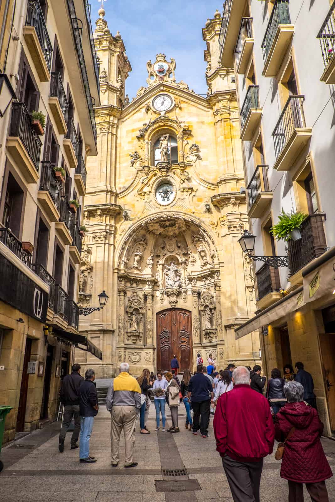 People walking down a street in San Sebastian towards the detailed facade of the Santa Maria Basilica.