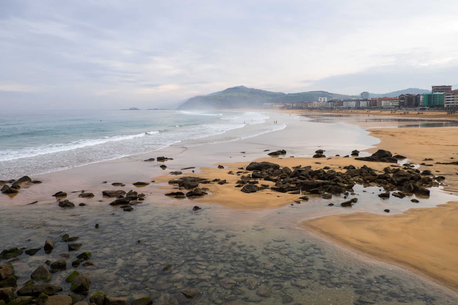 A wide stretch of yellow sand coastline strewn with rocks, backed by modern city buildings and low hills. 