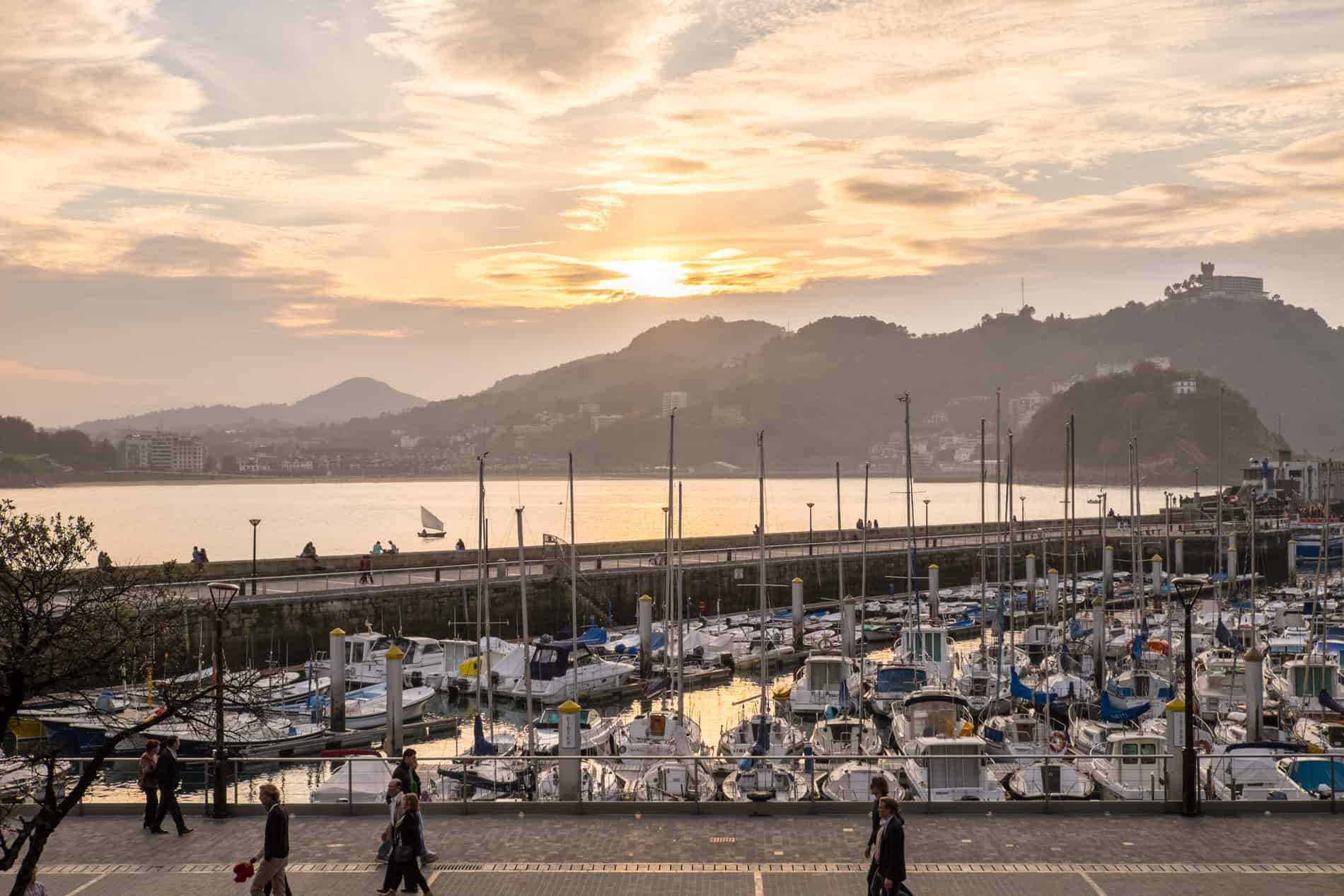 People on the promenade at sunset, next to the hilly port filled with white boats in San Sebastian city. 
