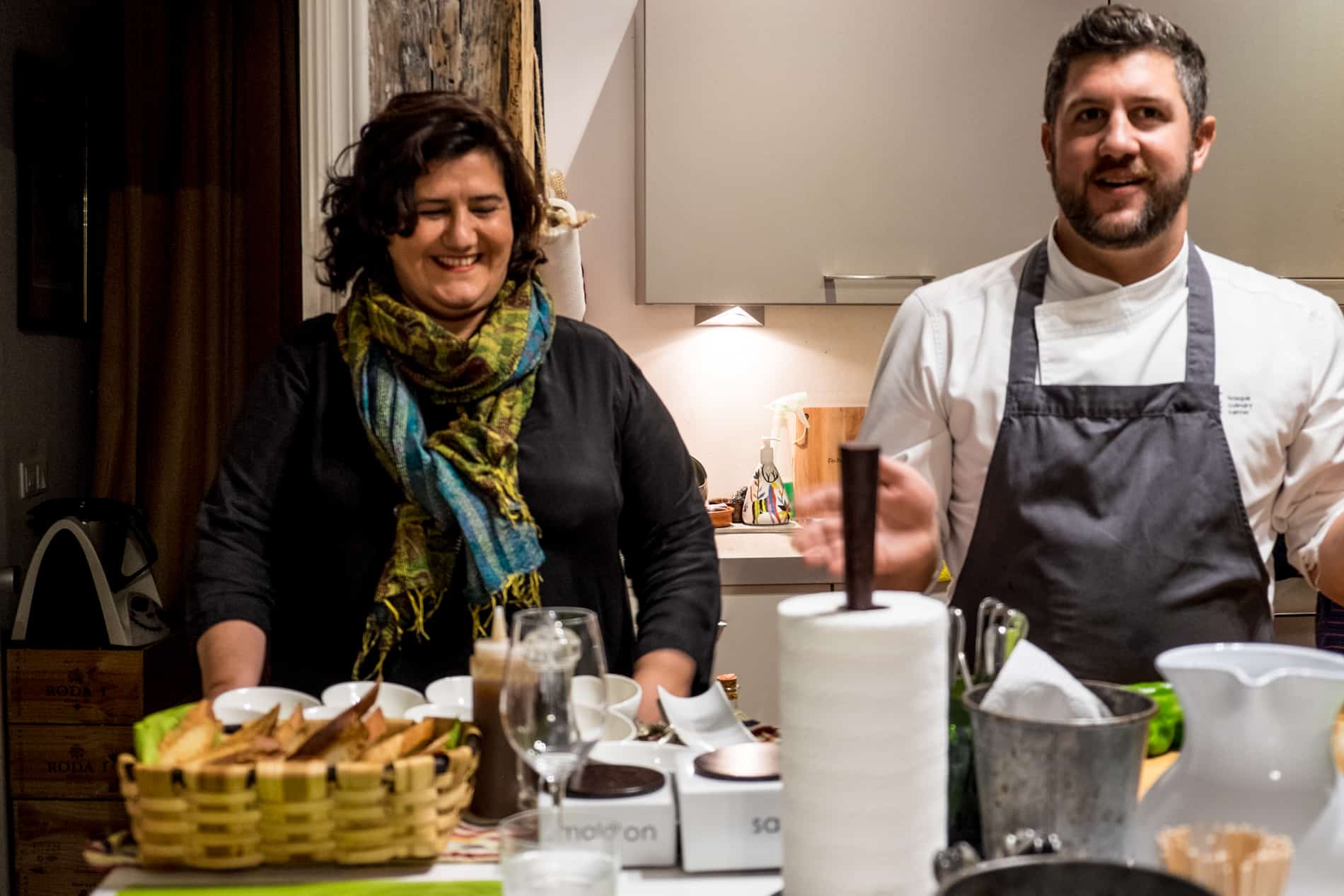 In a small kitchen, a woman stands with a chefs in whites and a grey apron, leading a cooking class. 