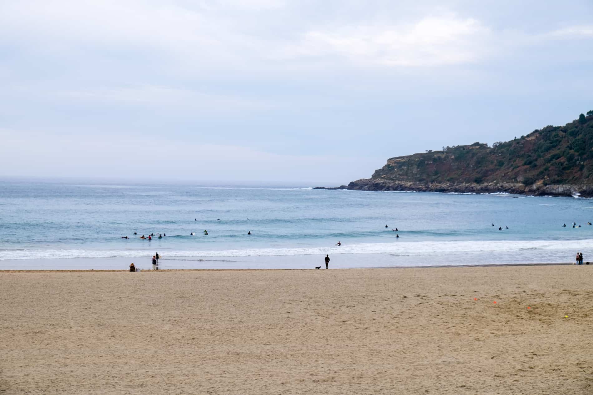 People on a yellow sandy beach watching the surfers out at sea. 