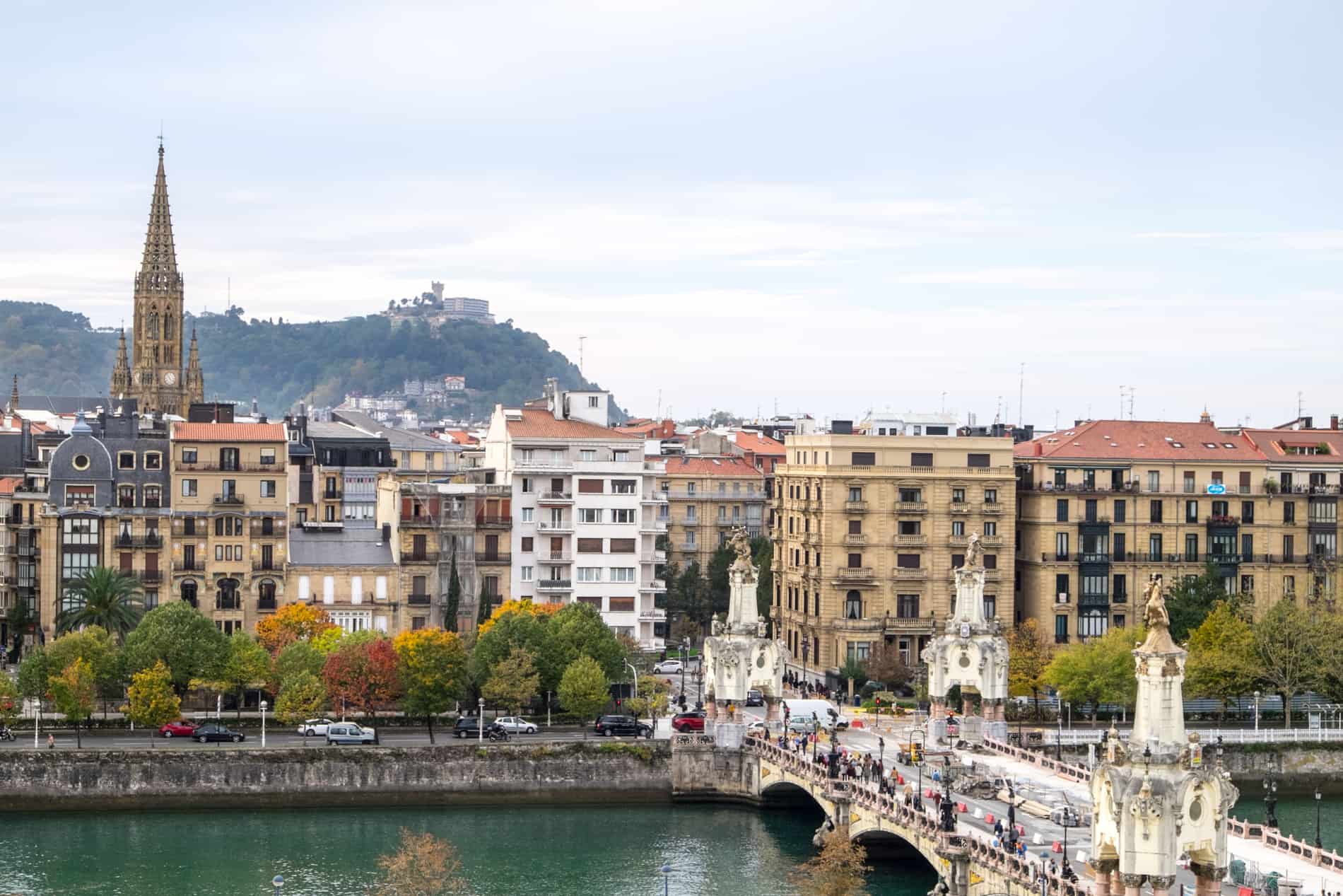 A white bridge over a emerald river leading to a town with caramel and white apartments buildings and a church spire, backed by a hill. 