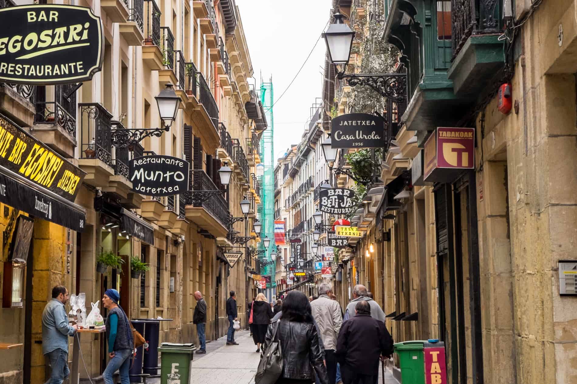 People walking down Fermin Calbeton Street - San Sebastian's food street lined with restaurant and Pintxos bar signs.