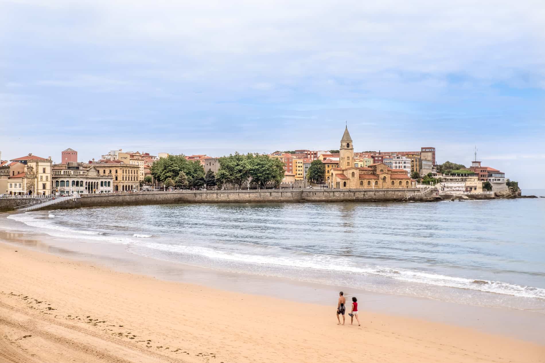 Two people walk along the beach in front of the old town of Gijón, whose pastel coloured buildings stand next to the sea. 