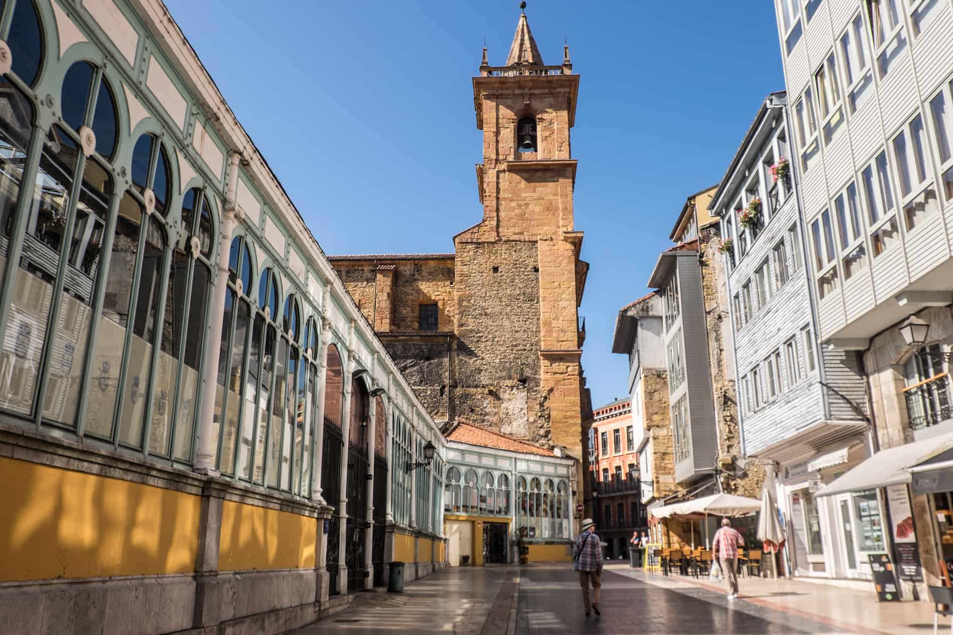 Two men walk down a street towards an old stone church, with a covered market to the left and a white wooden facade building to the right. 