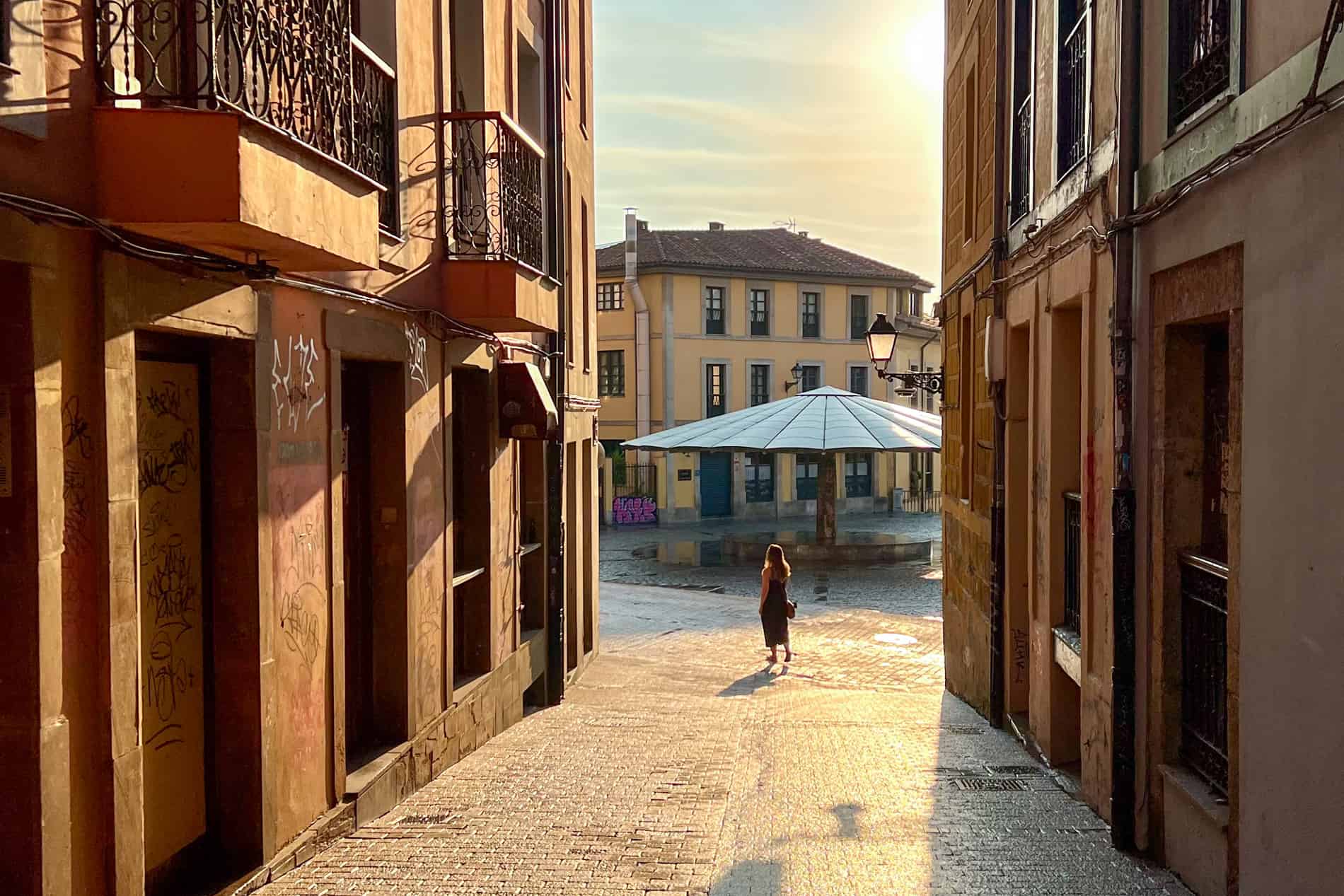 A woman walks down a street in Oviedo during morning golden hour, towards a giant umbrella structure in a public square.