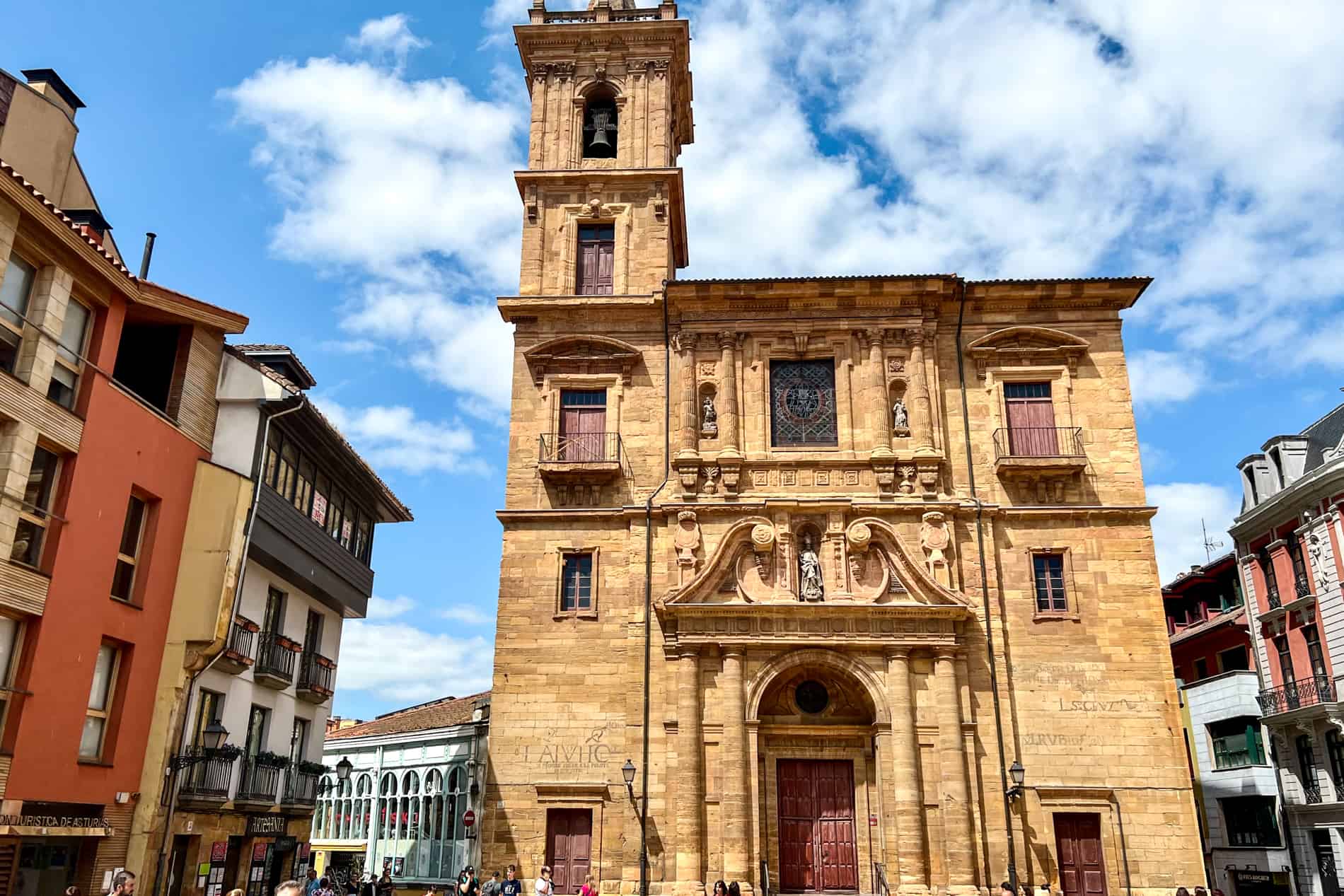 A caramel brown stone square church building with a single tower and rusty red window shutters in Oviedo, Spain. 