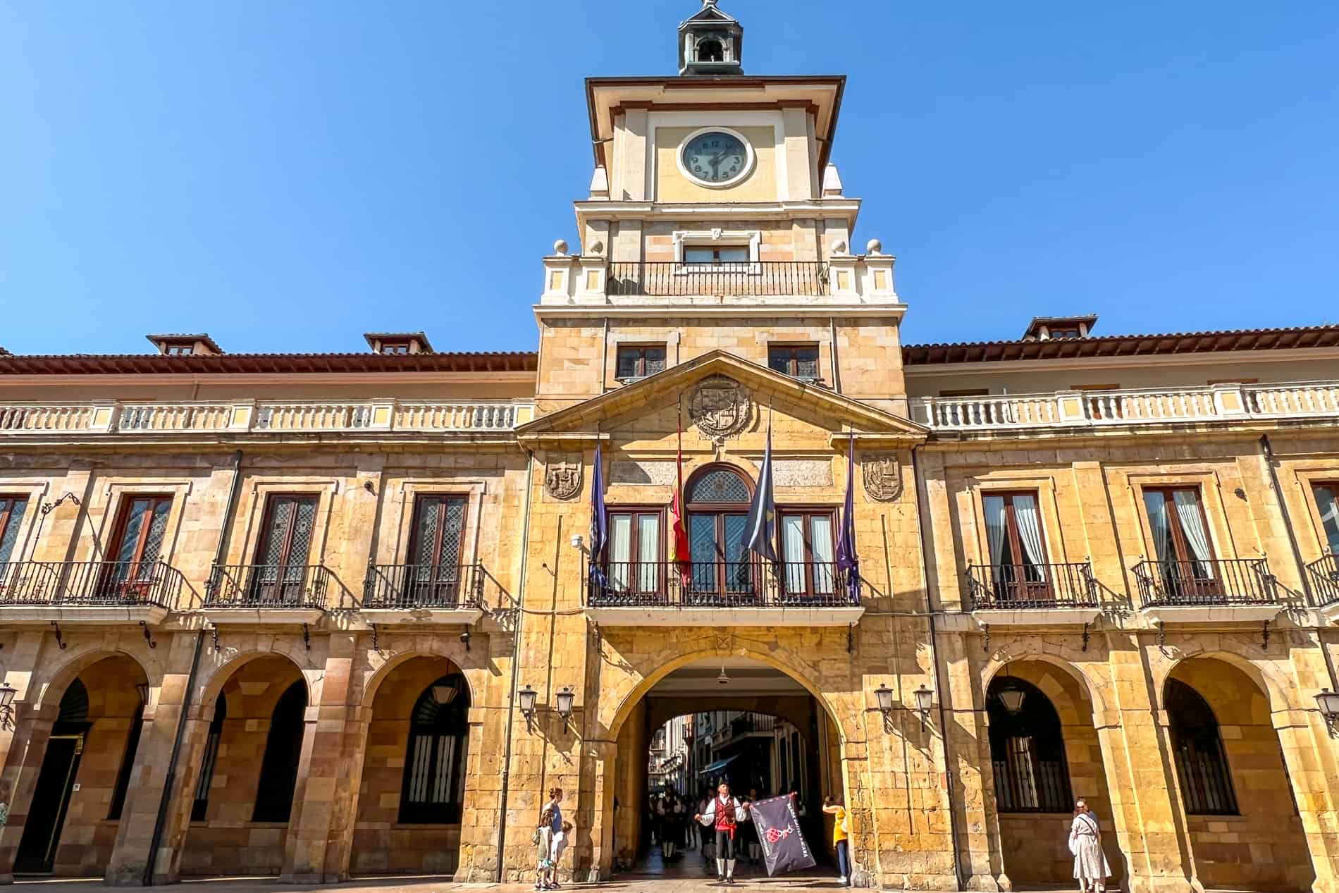 A long golden stone building with central clock tower is Oviedo's Town Hall. A man in traditional dress, holding a flag, stands in the archway. 