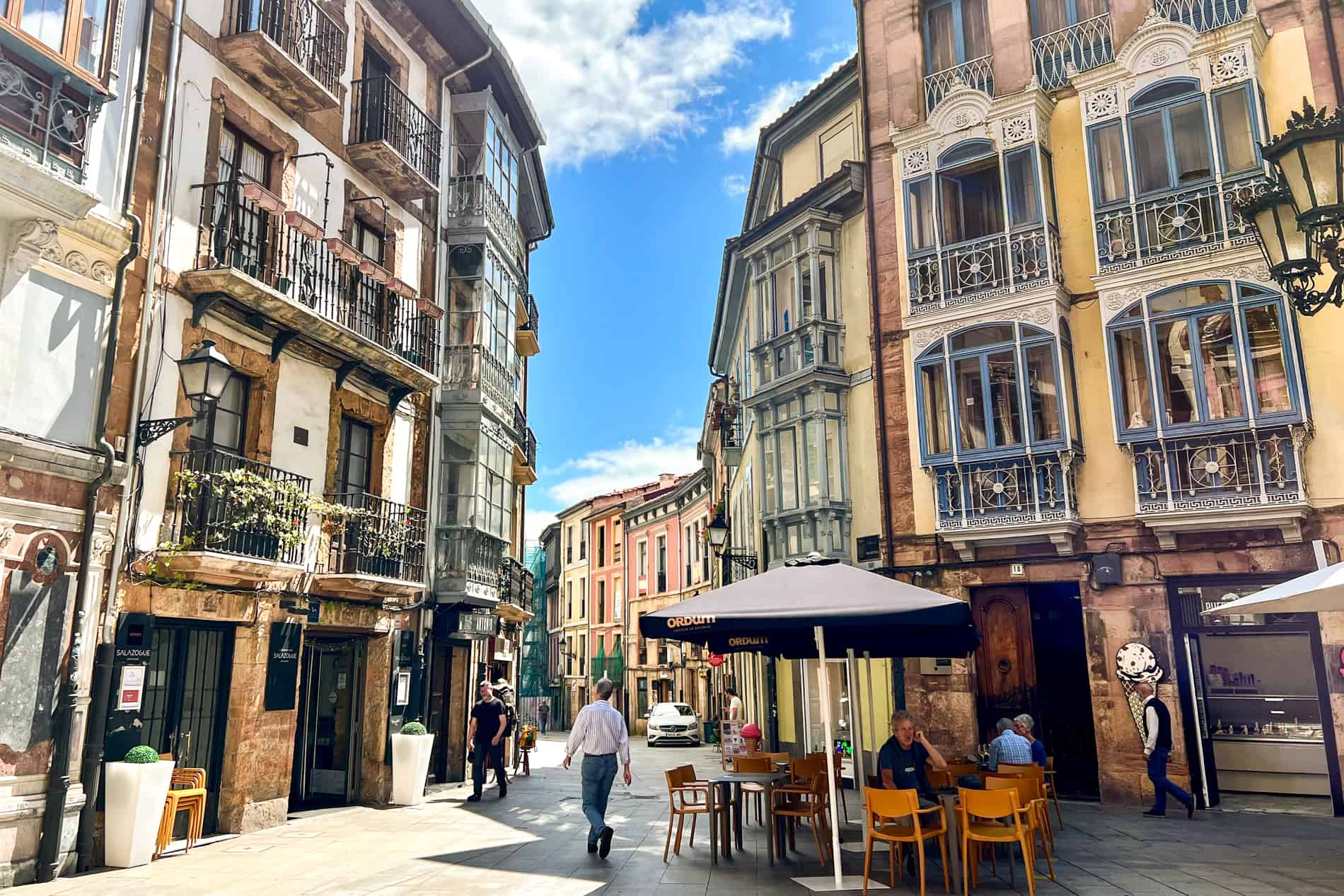 People sit outside at a cafe amongst pretty, pastel painting buildings with iron balconies on a street in Oviedo, Spain. 
